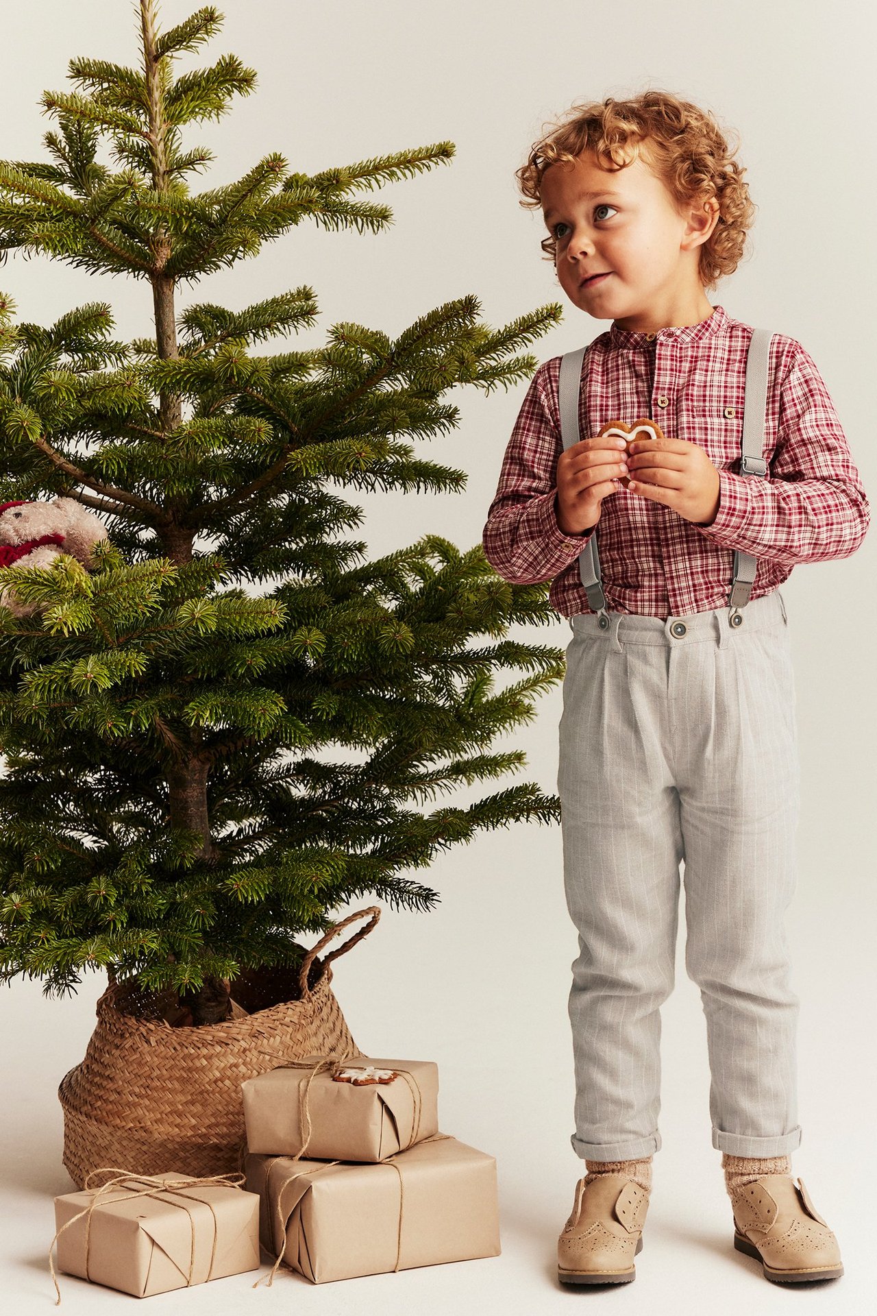Front view, kid in beige pinstripe suspender trousers, red plaid shirt, beige shoes, holding cookie.
