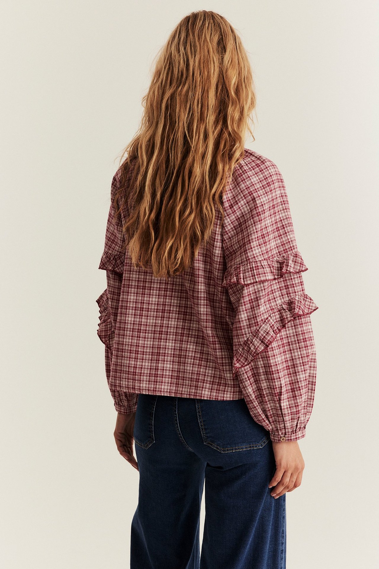 Back view of woman in red plaid woven blouse with ruffled sleeves, paired with dark blue jeans.