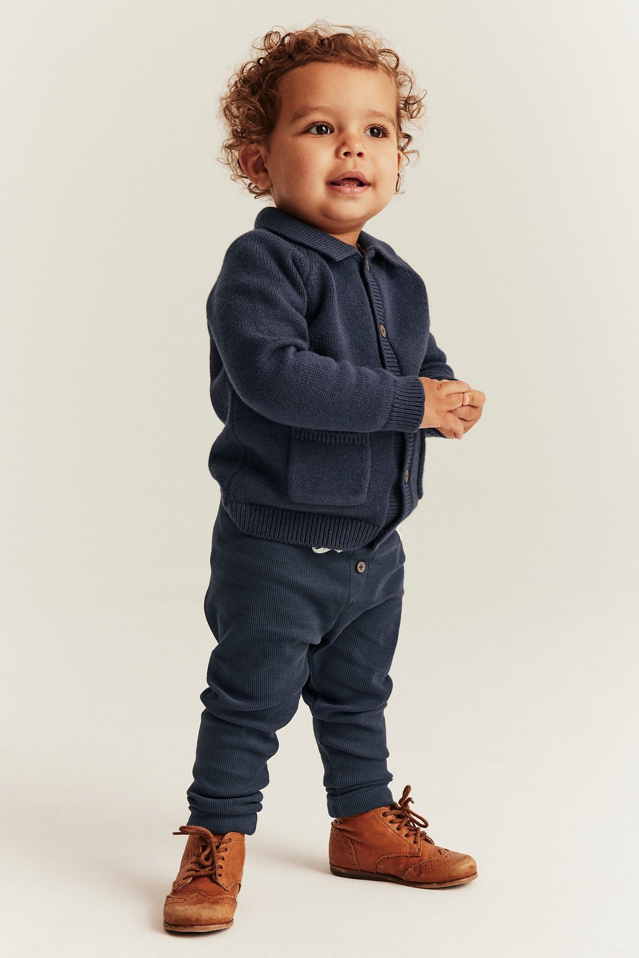 Front view of a baby wearing dark blue ribbed leggings, a matching knitted cardigan, and brown lace-up shoes.