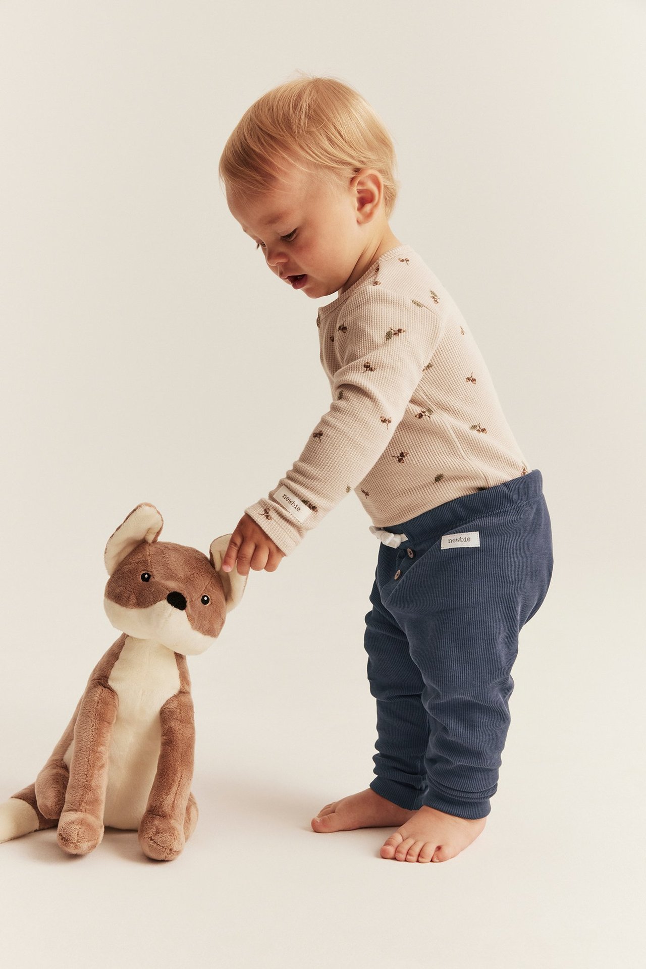 Side view of a baby in dark blue ribbed leggings with a drawstring and a beige patterned top, touching a fox toy.