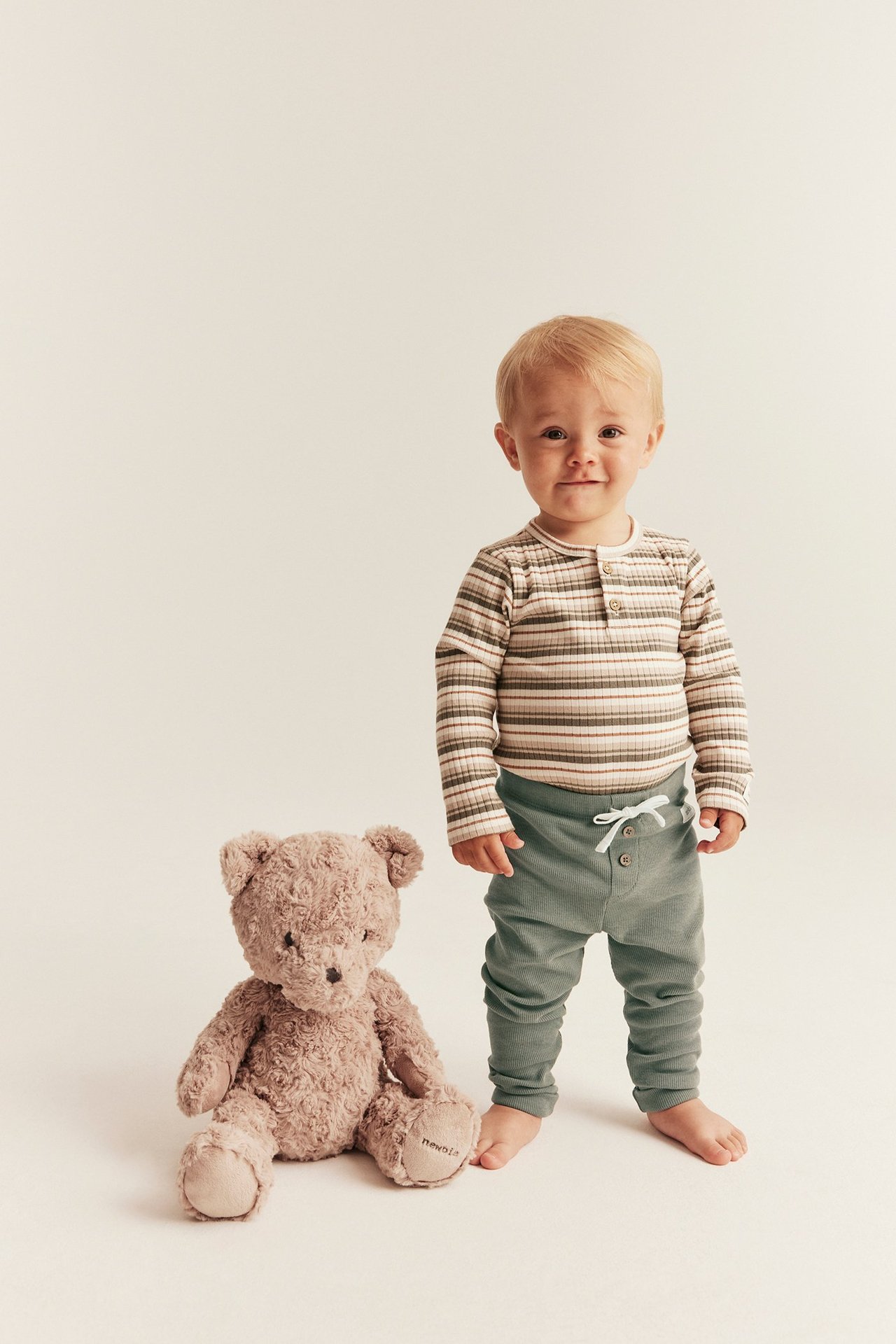 Frontansicht: Baby in dunkelgrünen gerippten Leggings, gestreiftem Oberteil, neben einem braunen Teddybär.