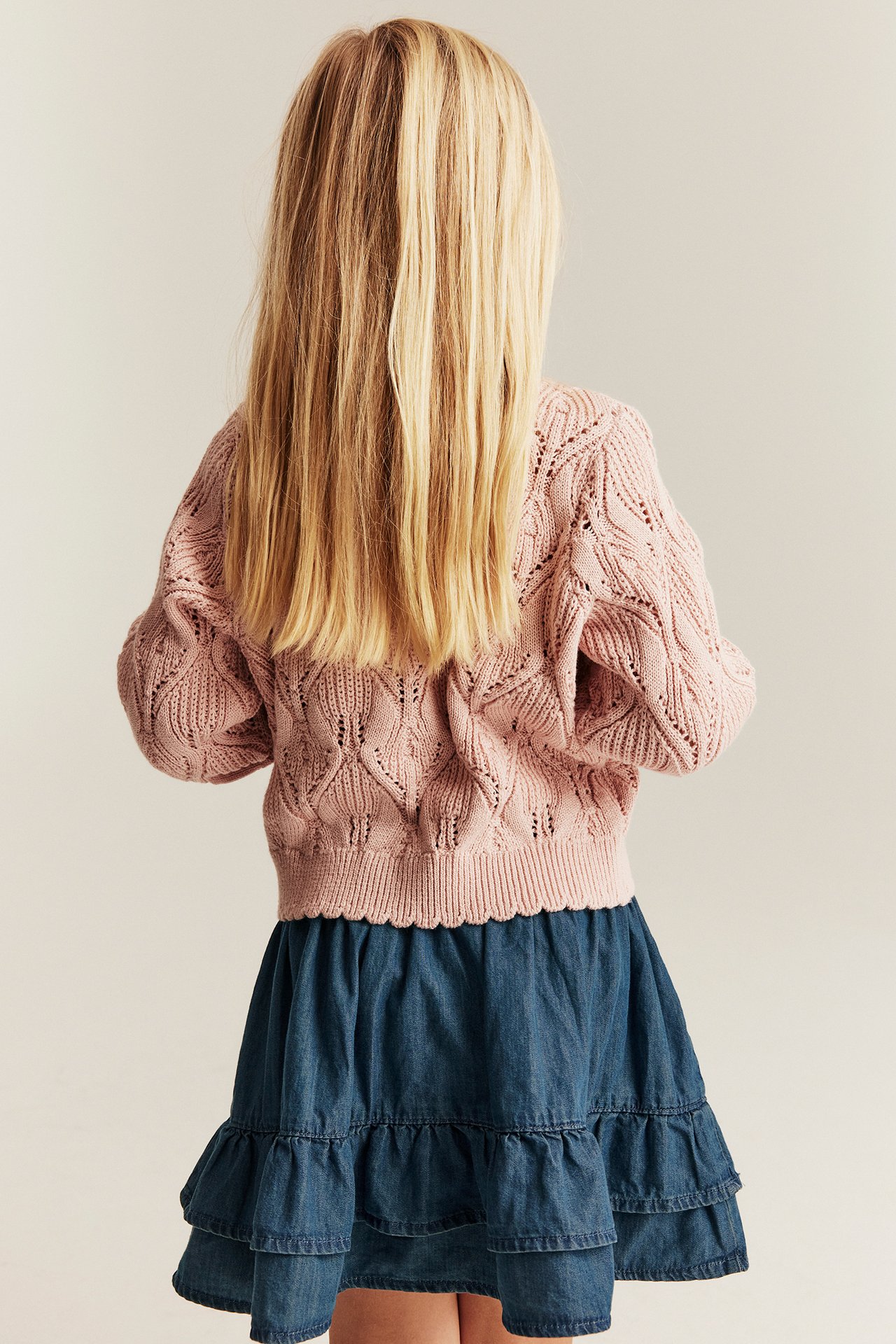 Rückansicht eines Kindes in blauem Jeansrock mit Rüschen und hellrosa Strickpullover.
