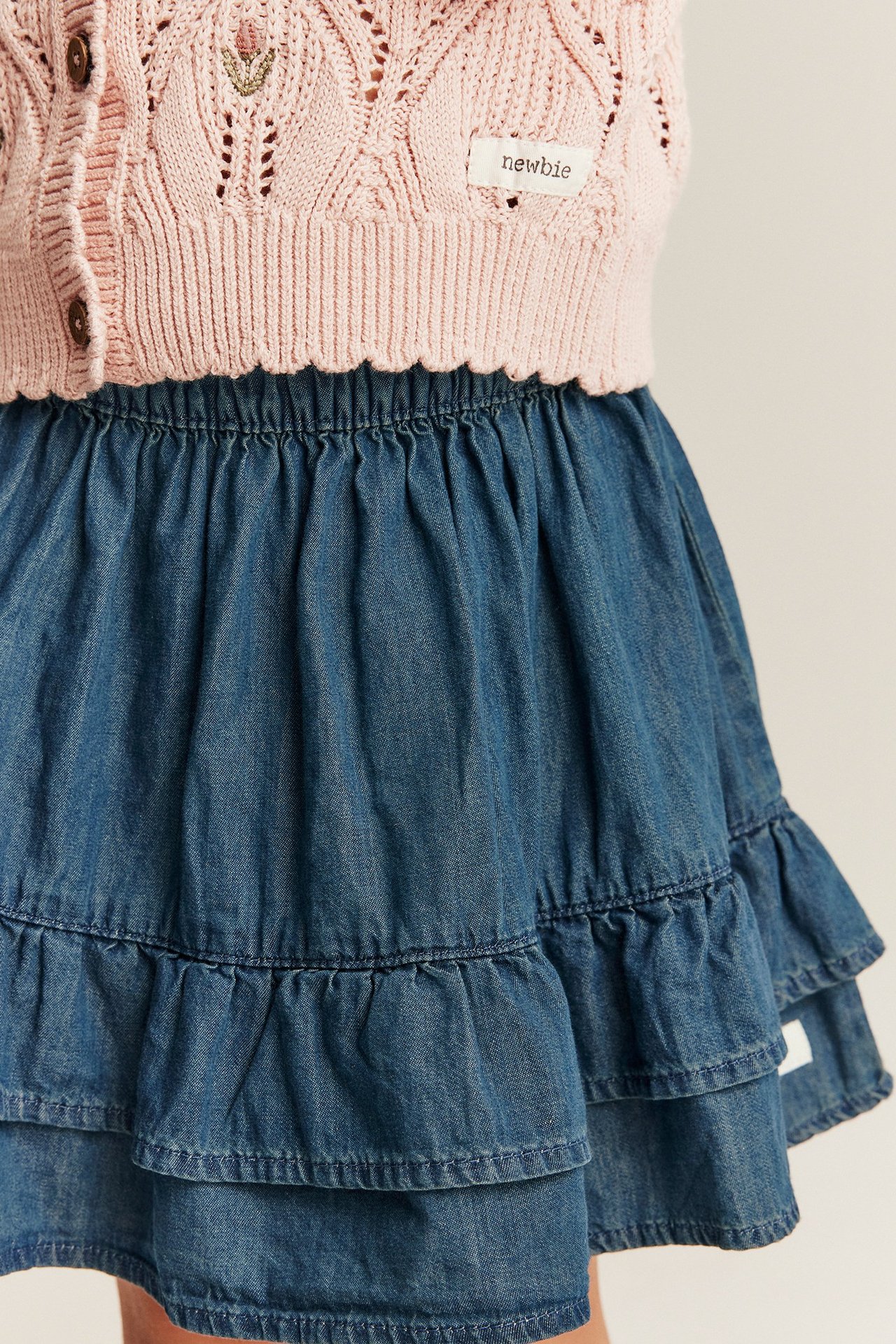 Nahaufnahme eines Kinder-Denimrocks in Blau mit Gummibund, Rüschen und einer rosa Strickjacke.