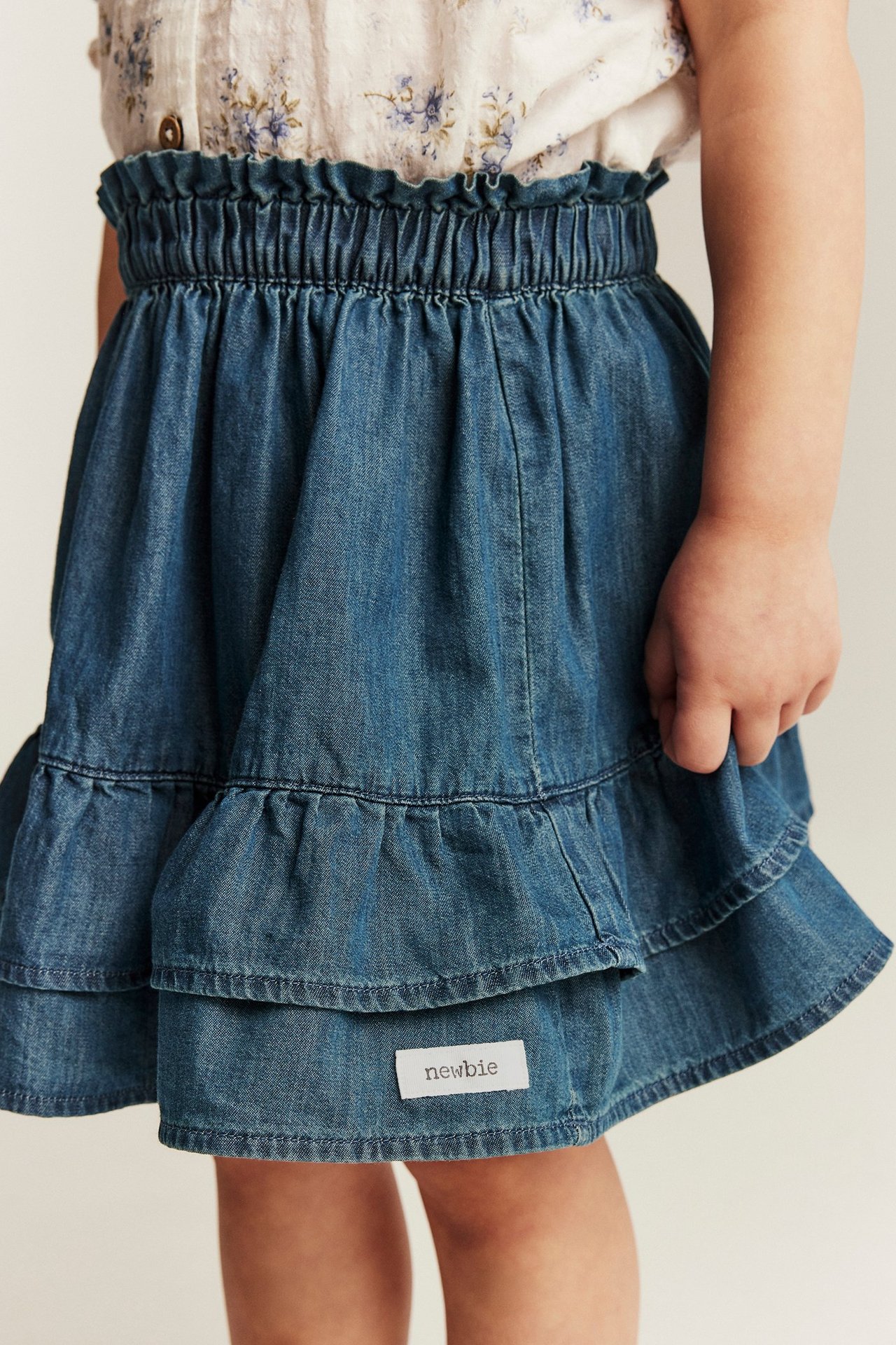 Close-up front view of a kid's blue denim paperbag skirt with ruffled hem over a floral shirt.