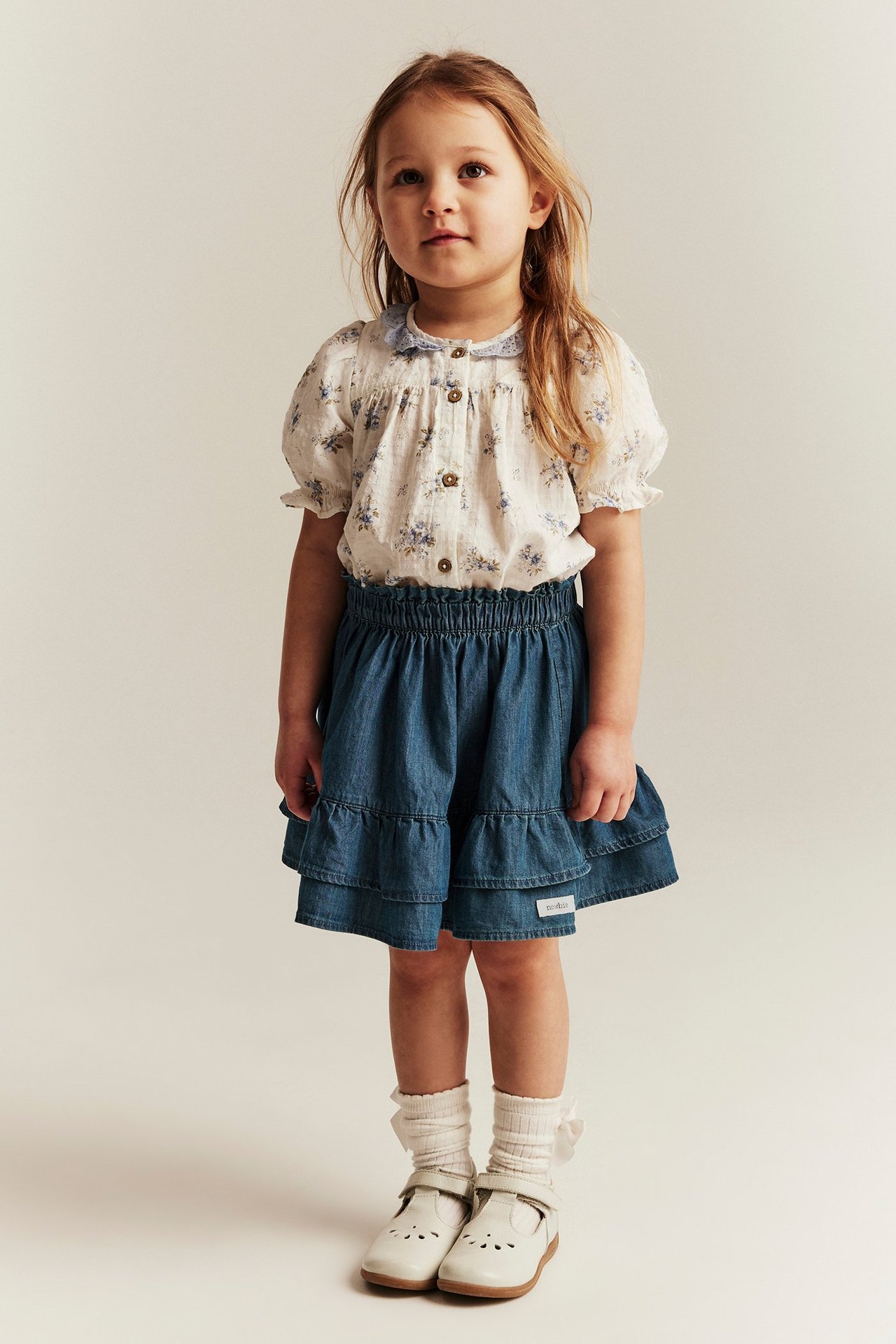 Front view: Kid in a blue ruffled denim skirt, white floral blouse, white socks, and white shoes.