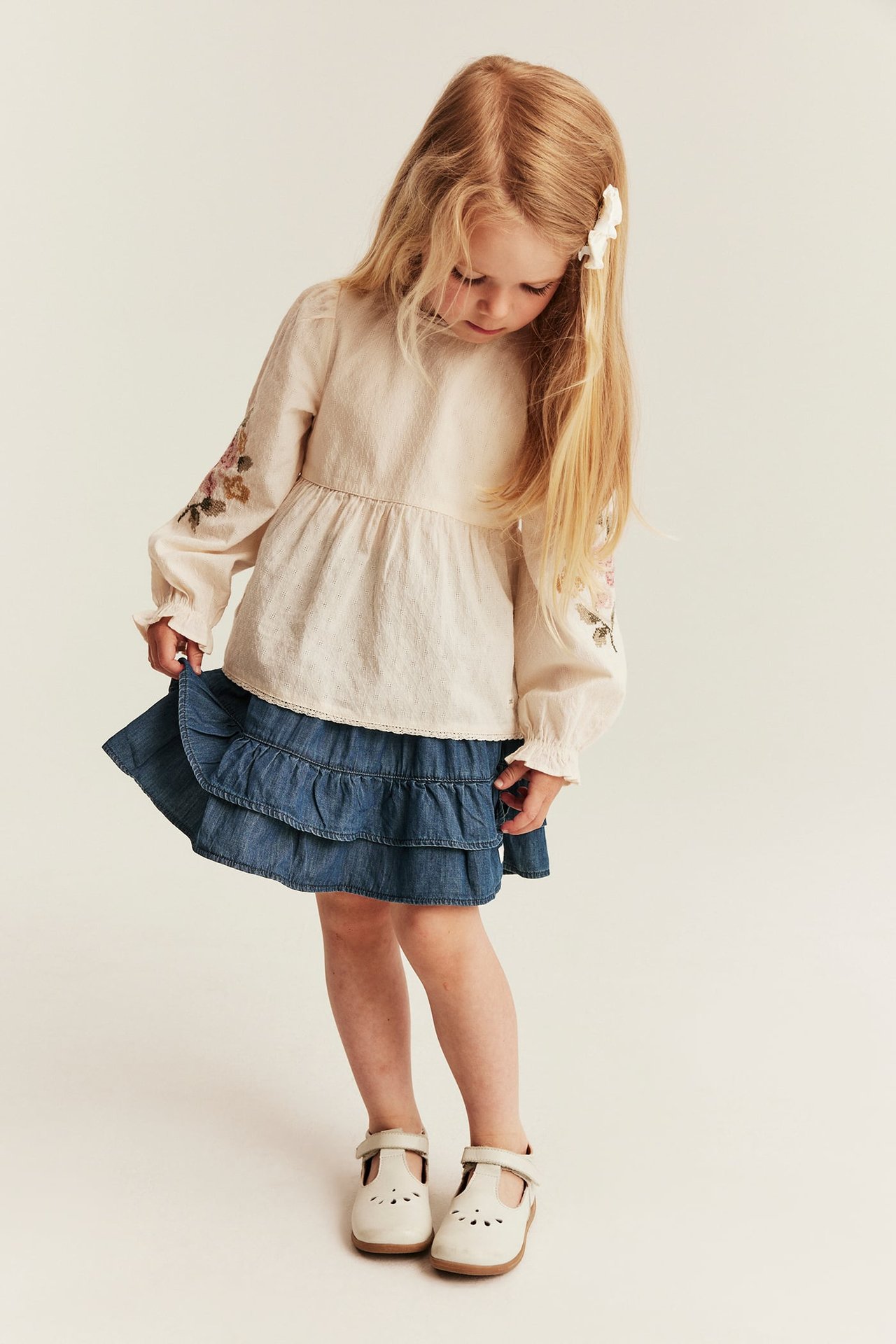 Vorderansicht: Kind in blauem Jeans-Volantrock, beiger bestickter Bluse, weißer Haarspange und T-Spangen-Schuhen.