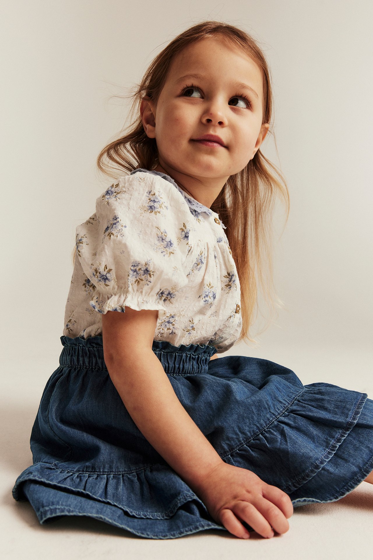 Front view of a kid sitting, wearing a blue denim ruffled skirt and a cream floral blouse.