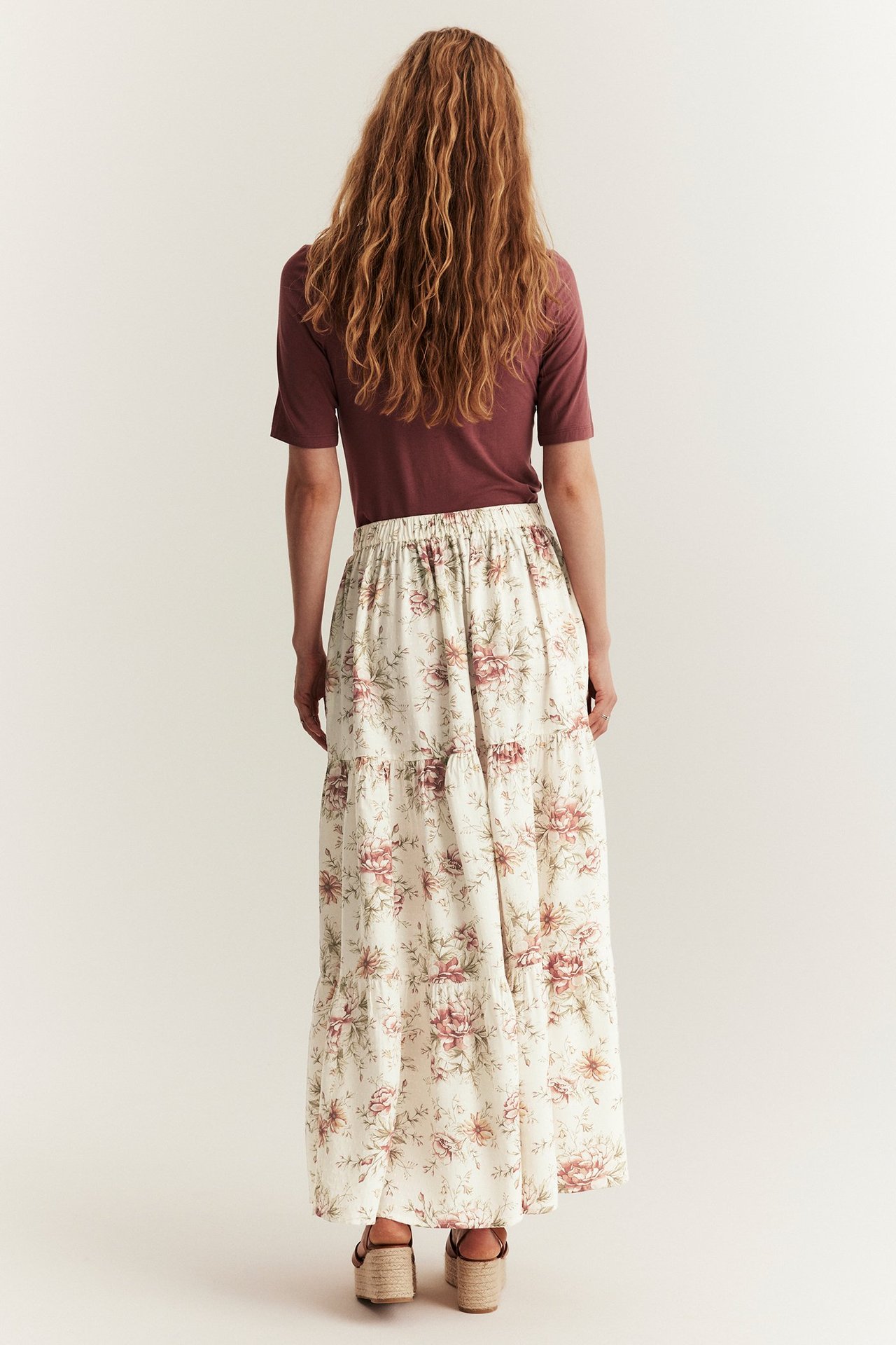 Back view: Woman in off-white floral tiered long skirt, brown short-sleeved top, and wedge sandals.