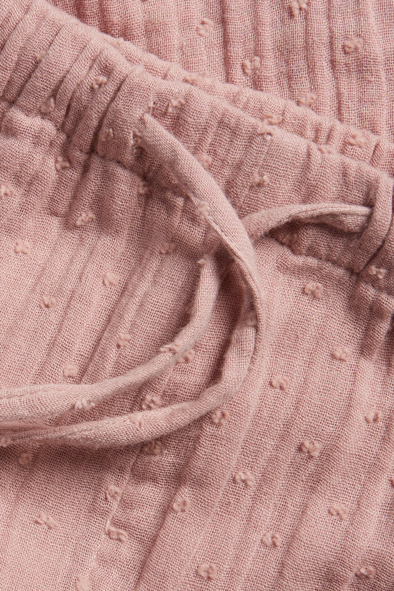 Close-up of pink woven cotton trousers with an elasticated waist and drawstring, featuring a dotted texture.