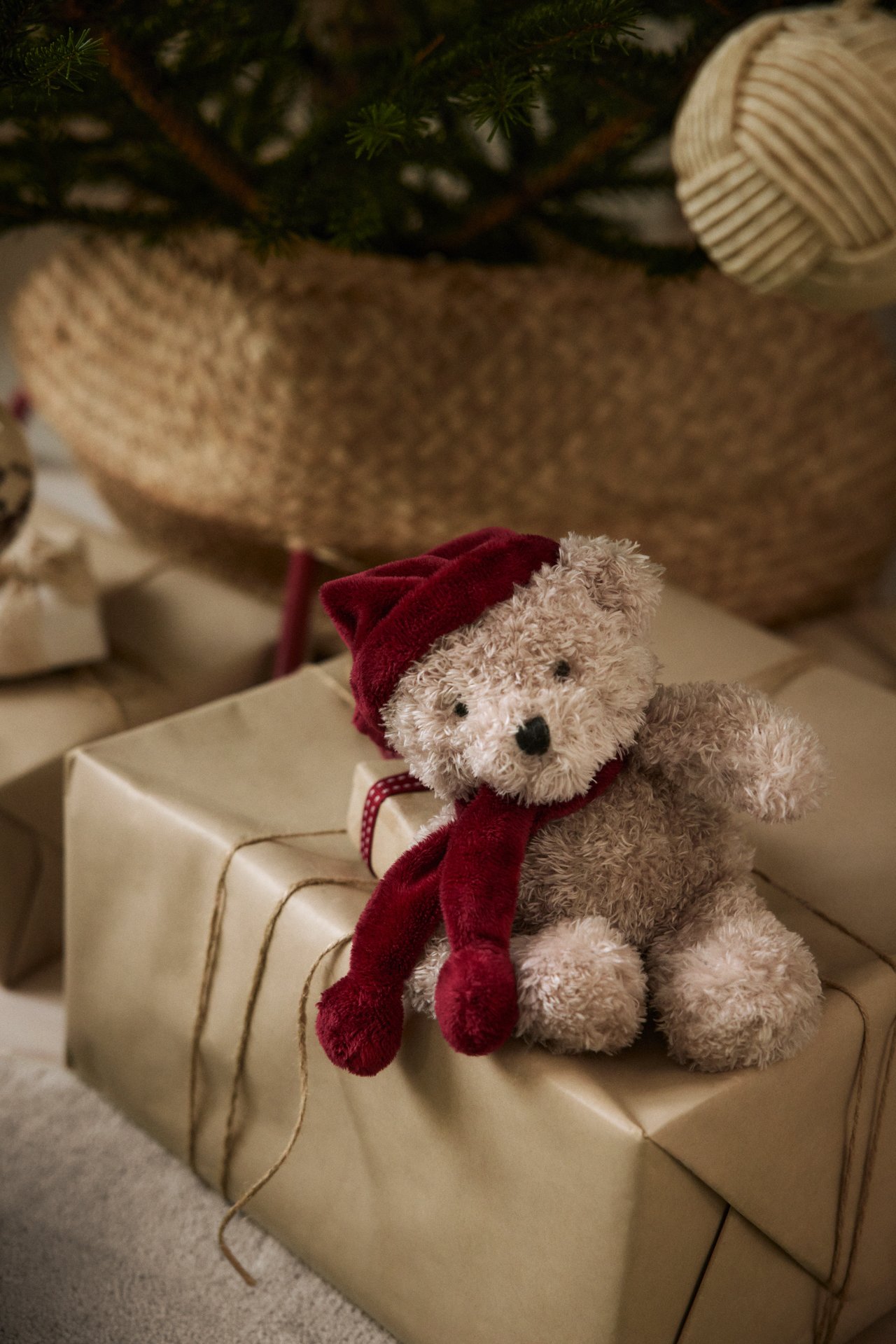 Nahaufnahme eines hellbraunen Teddybären mit roter Weihnachtsmütze und Schal, sitzend auf braunen Geschenken.