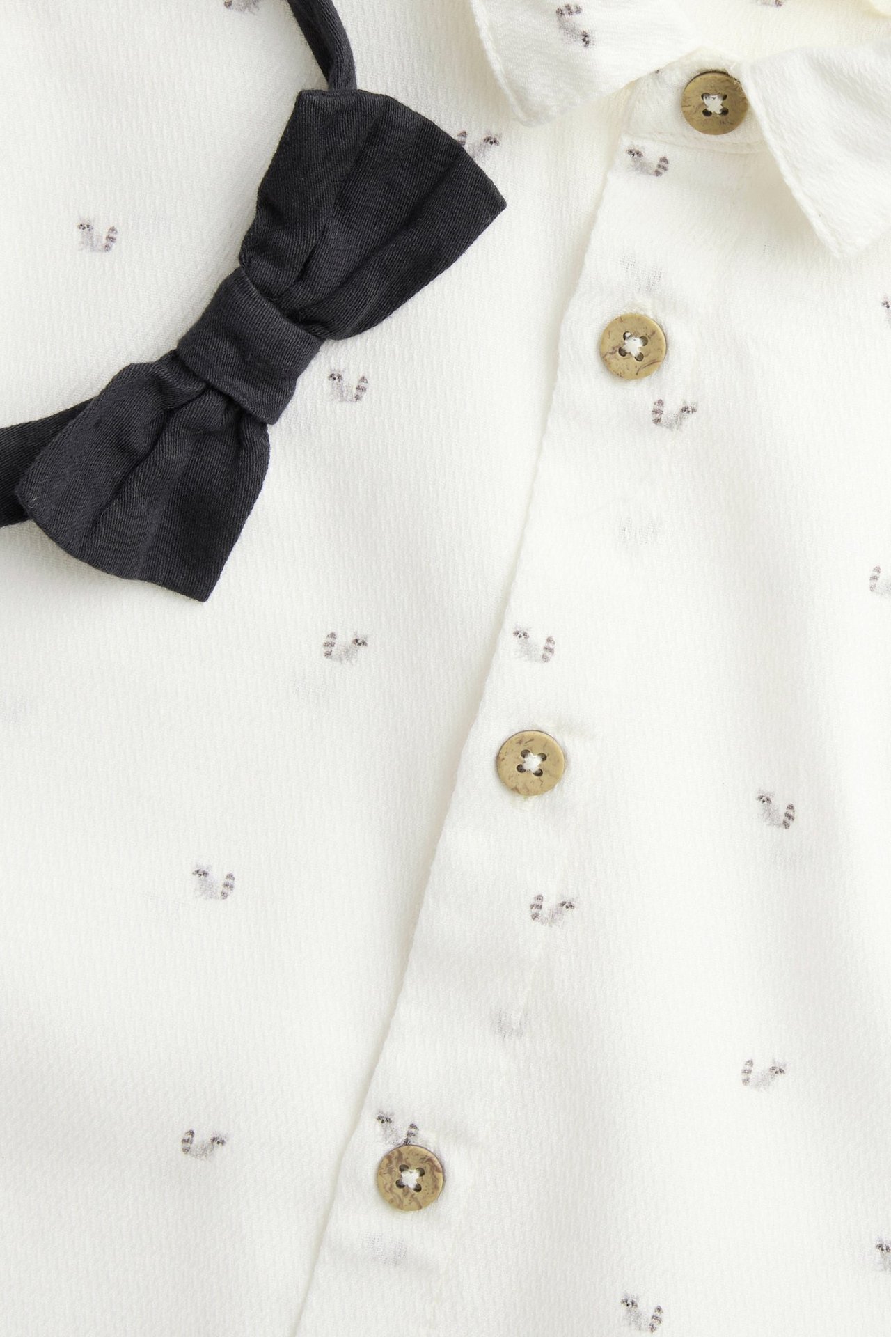 Close-up of an off-white woven dress shirt with animal pattern, brown buttons, and a black detachable bow tie.