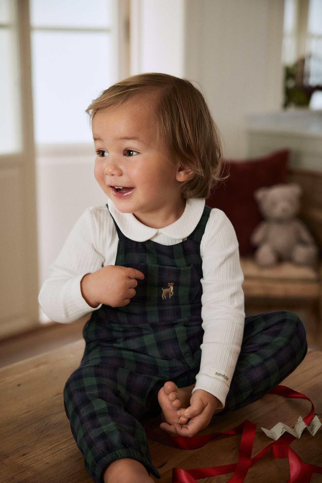 Front view: Baby in green plaid cotton dungarees with deer embroidery over a white ribbed top.