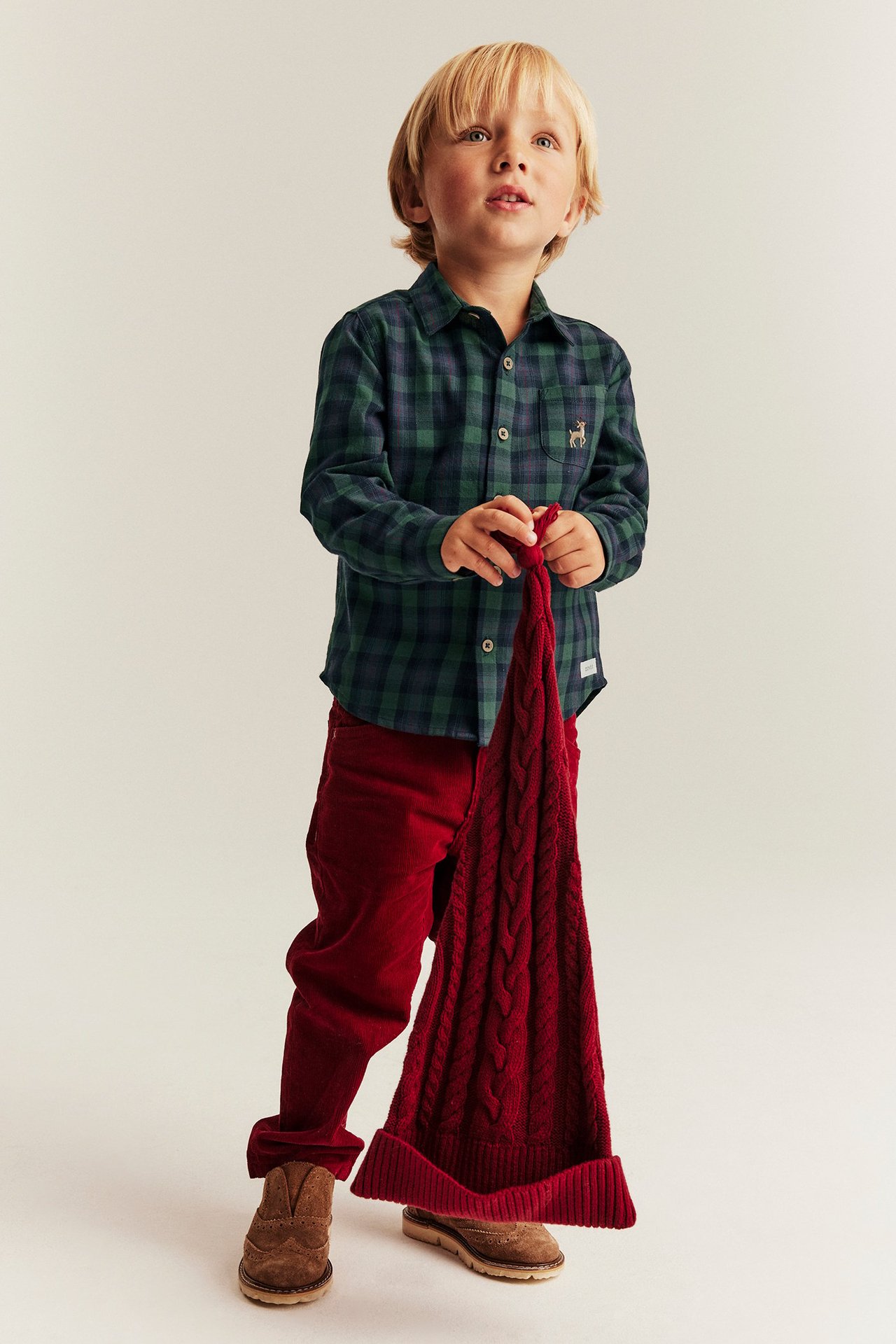 Mädchen in blauer karierten Weihnachtsbluse mit Kragen, rote Hose, hält rote Strickdecke.