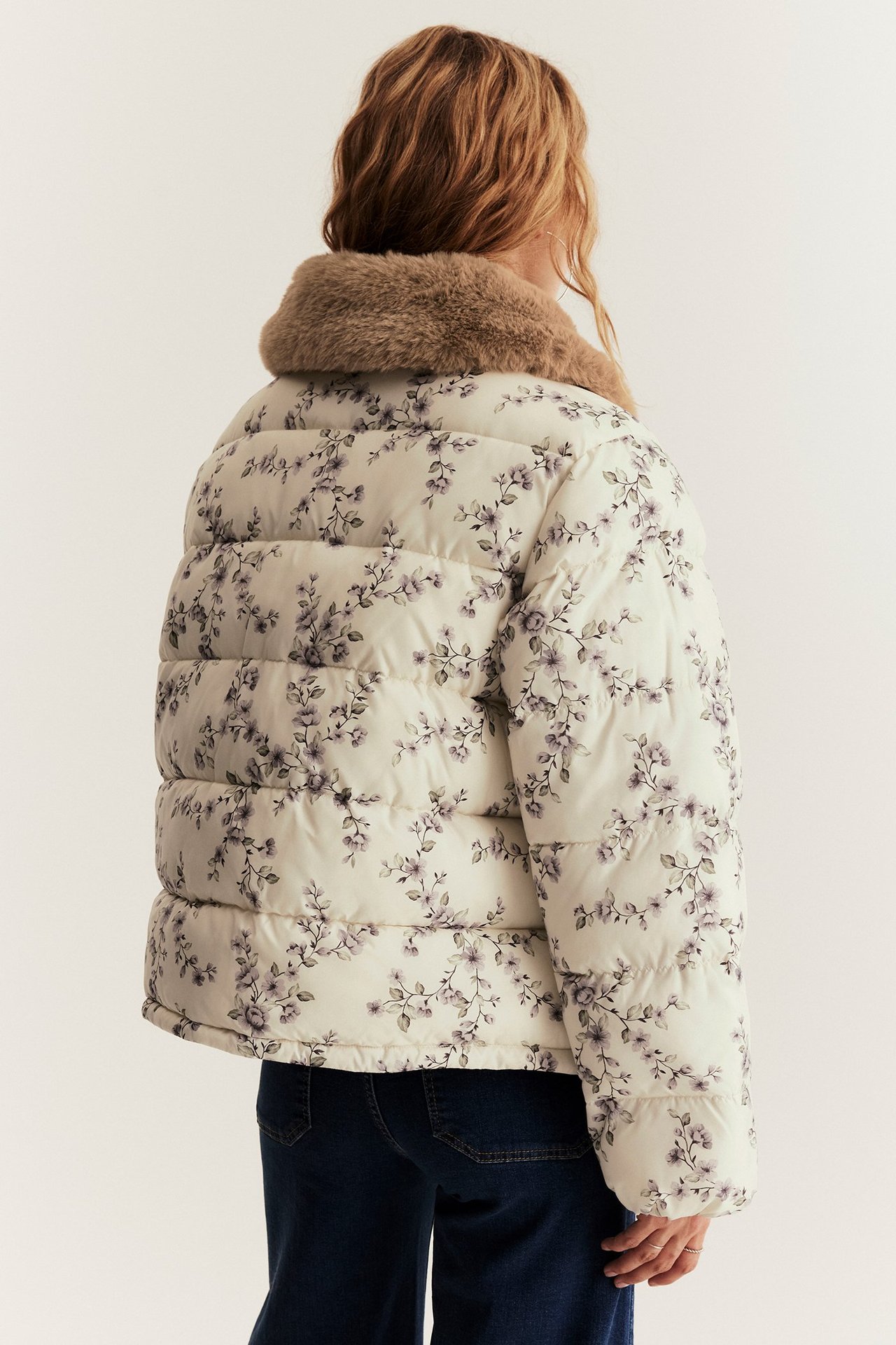 Back view of a woman wearing a light beige floral padded jacket with a brown faux fur collar and dark blue jeans.