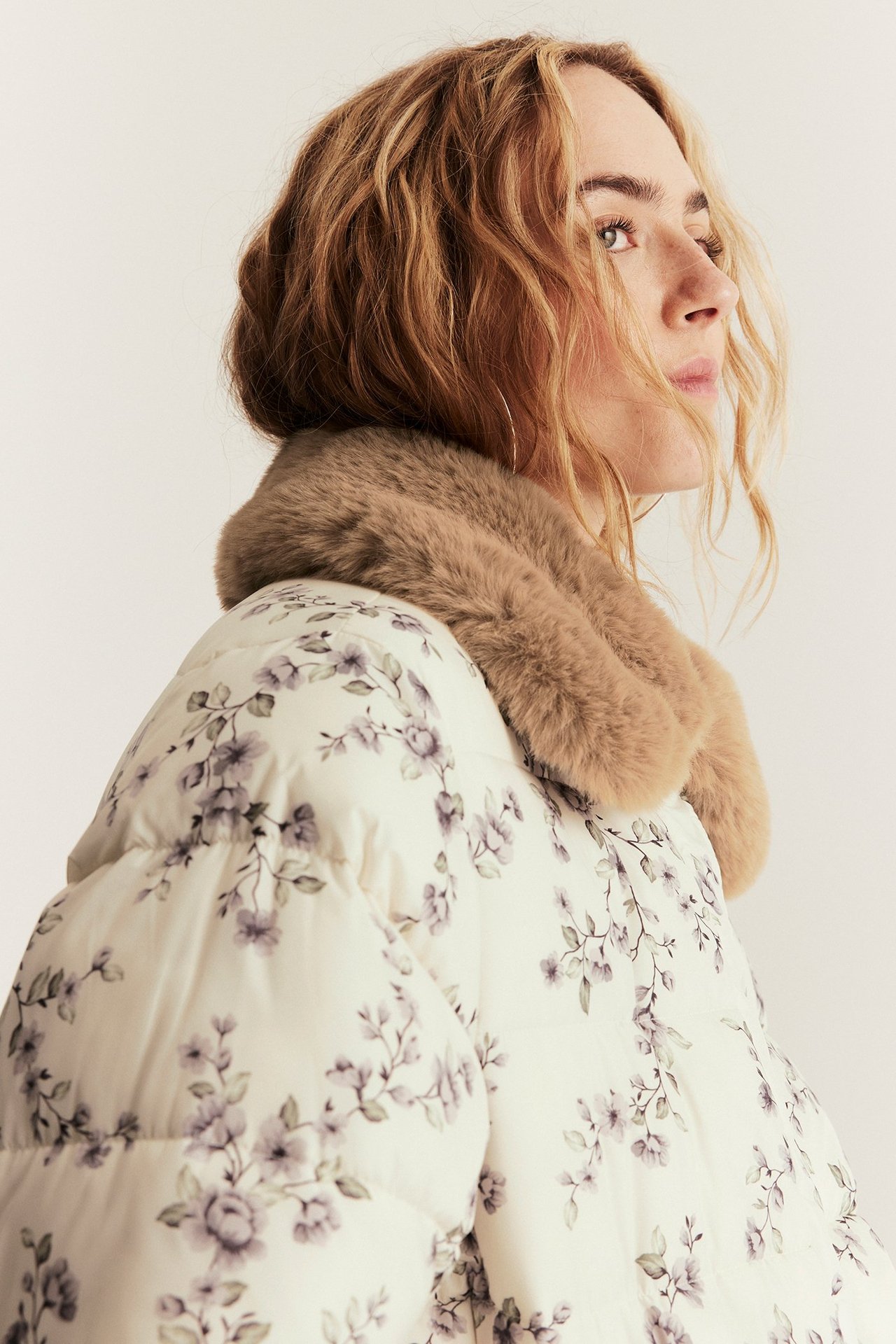 Side view of a woman wearing a light beige floral padded jacket with a brown faux fur collar and hoop earrings.