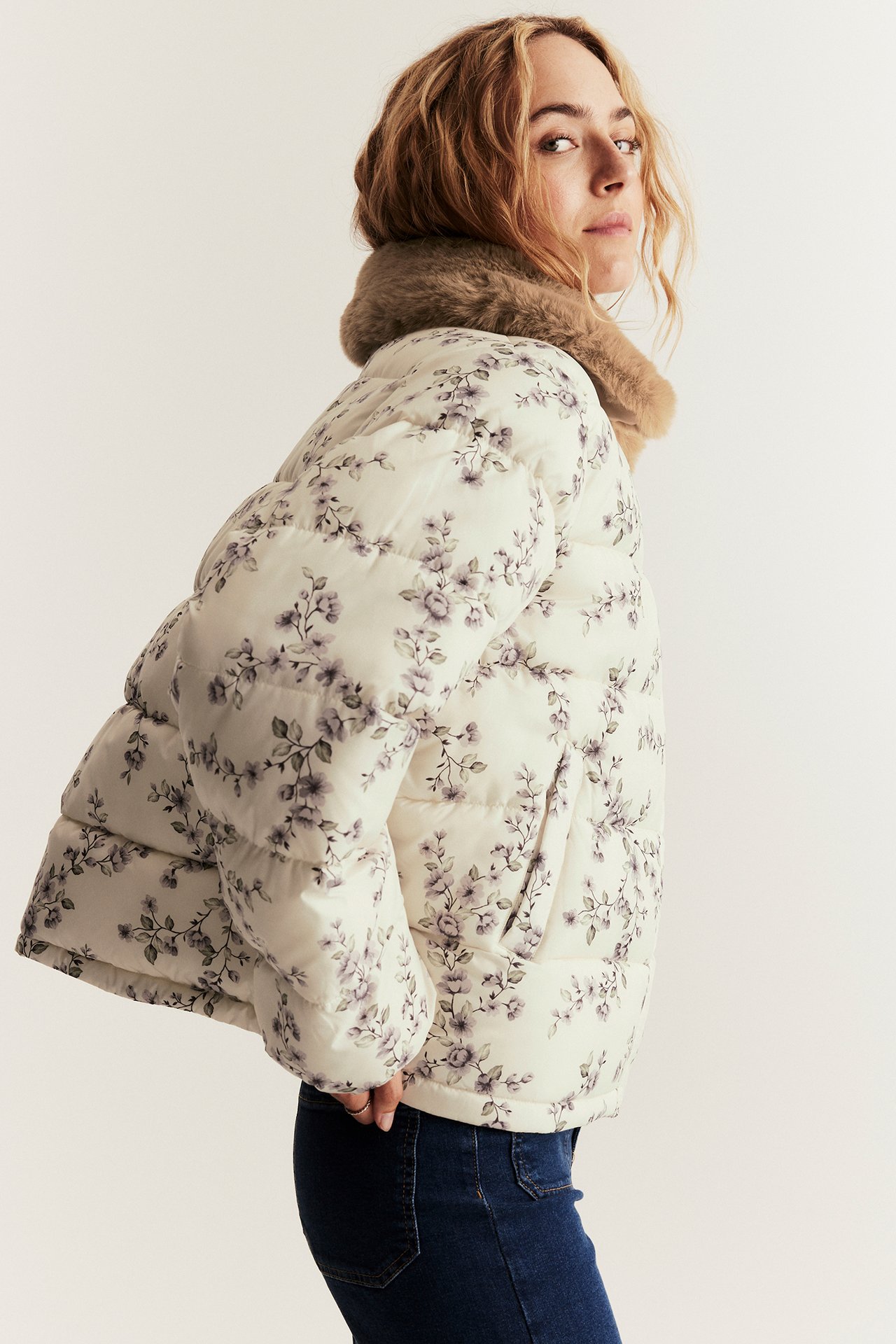 Side view: woman in light beige floral padded jacket with brown faux fur collar and dark blue jeans.