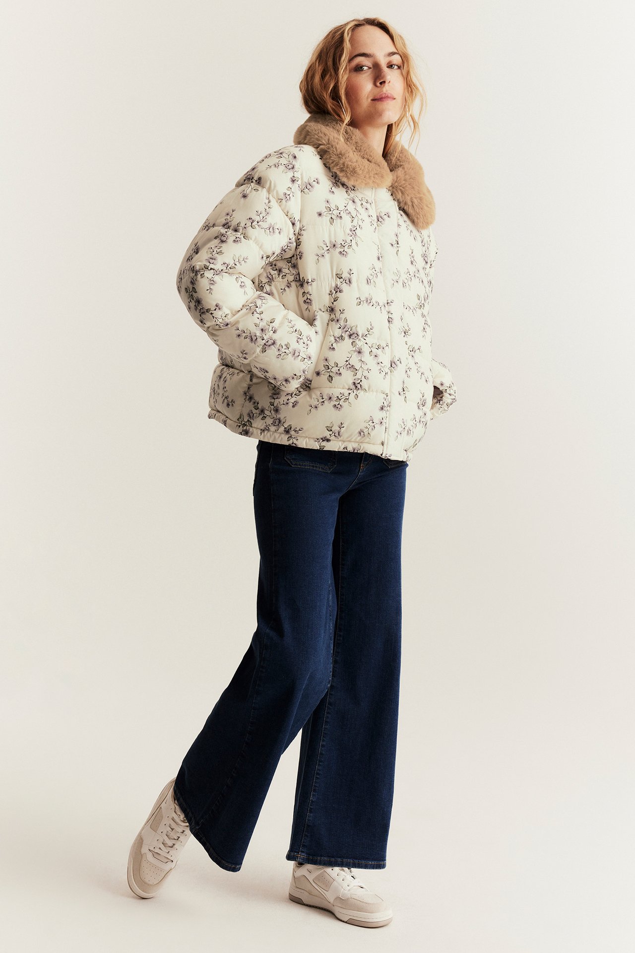 Front view: woman in light beige floral padded jacket with faux fur collar, dark blue jeans, and sneakers.
