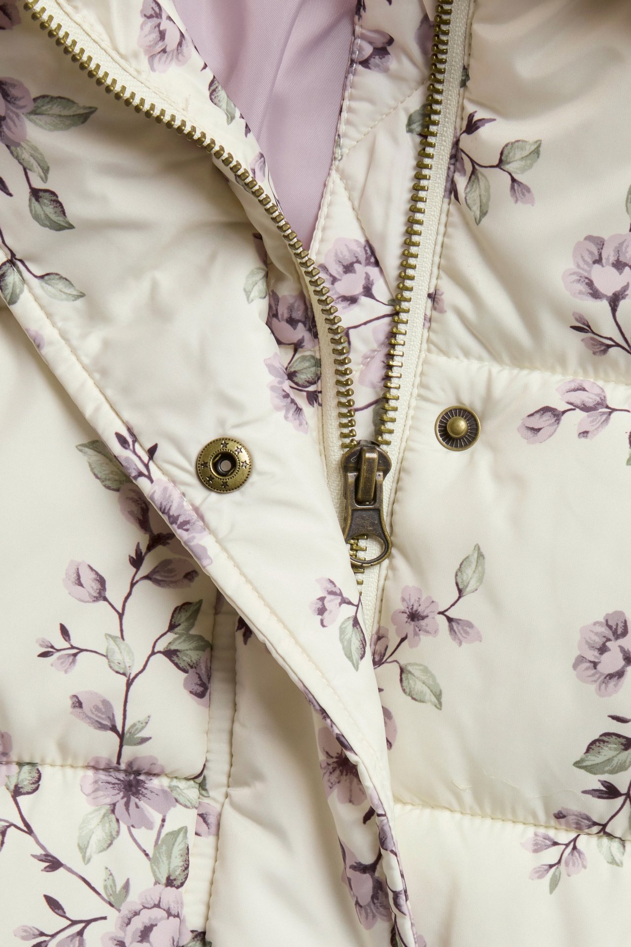 Nahaufnahme Vorderansicht: hellbeige wattierte Jacke mit lila Blumenmuster, Reißverschluss und Druckknöpfen.