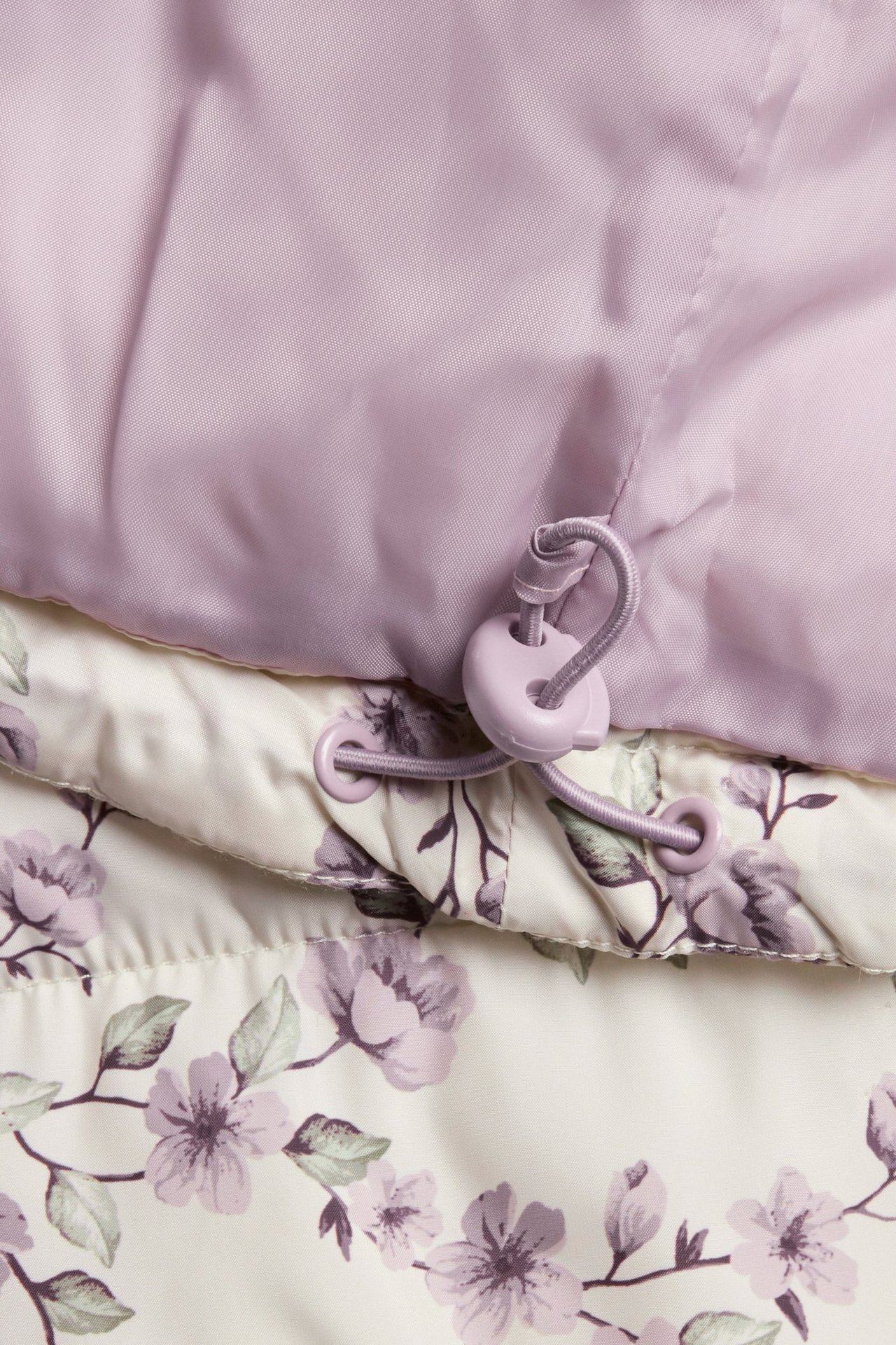 Close-up of a light beige padded jacket with a purple floral pattern and a lilac drawstring.