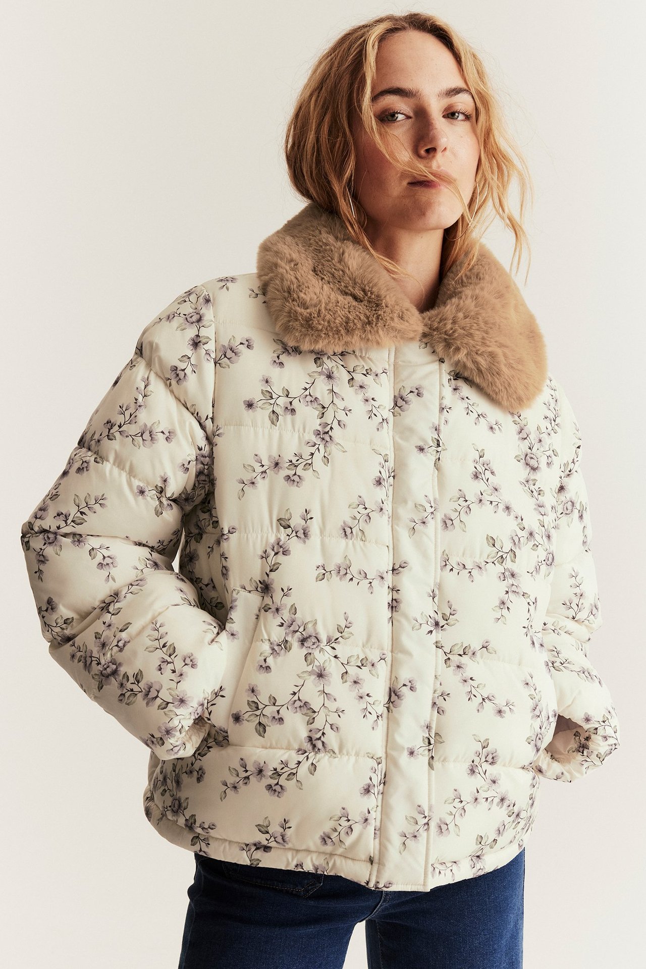 Front view of a woman wearing a light beige floral padded jacket with a brown faux fur collar and dark blue jeans.
