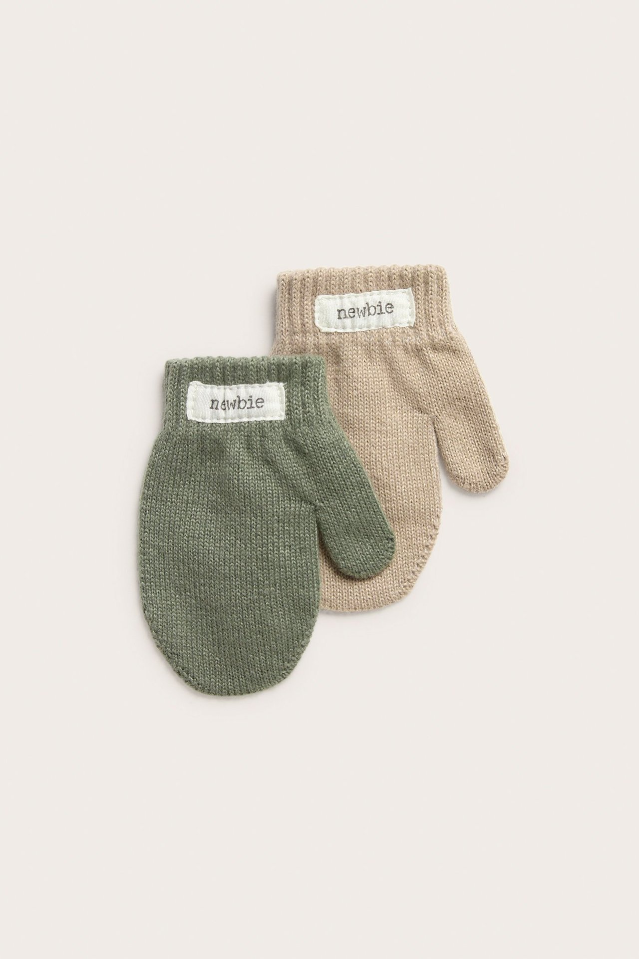 Draufsicht auf zwei gestrickte Babyfäustlinge, einen dunkelgrünen und einen beigen, mit Etiketten.