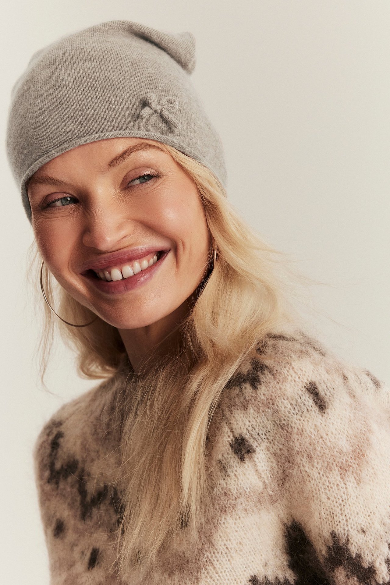 Front view: Woman wears beige melange wool-cashmere beanie with bow detail and patterned sweater.