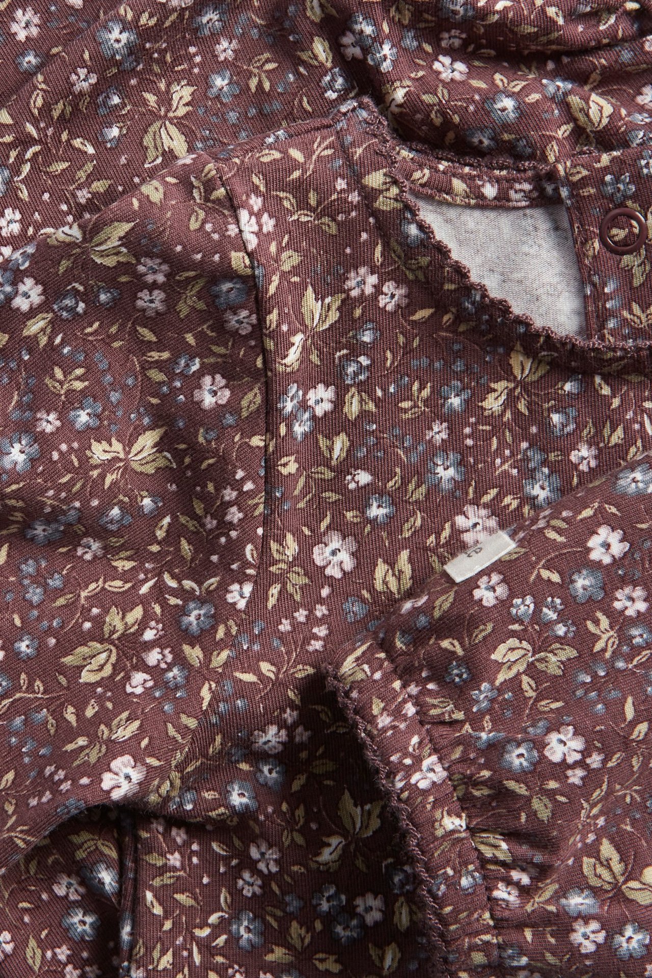 Nahaufnahme eines lila Baby-Bodys aus Baumwolle mit Blumenmuster, Puffärmeln, Picot-Borte und Druckknöpfen.