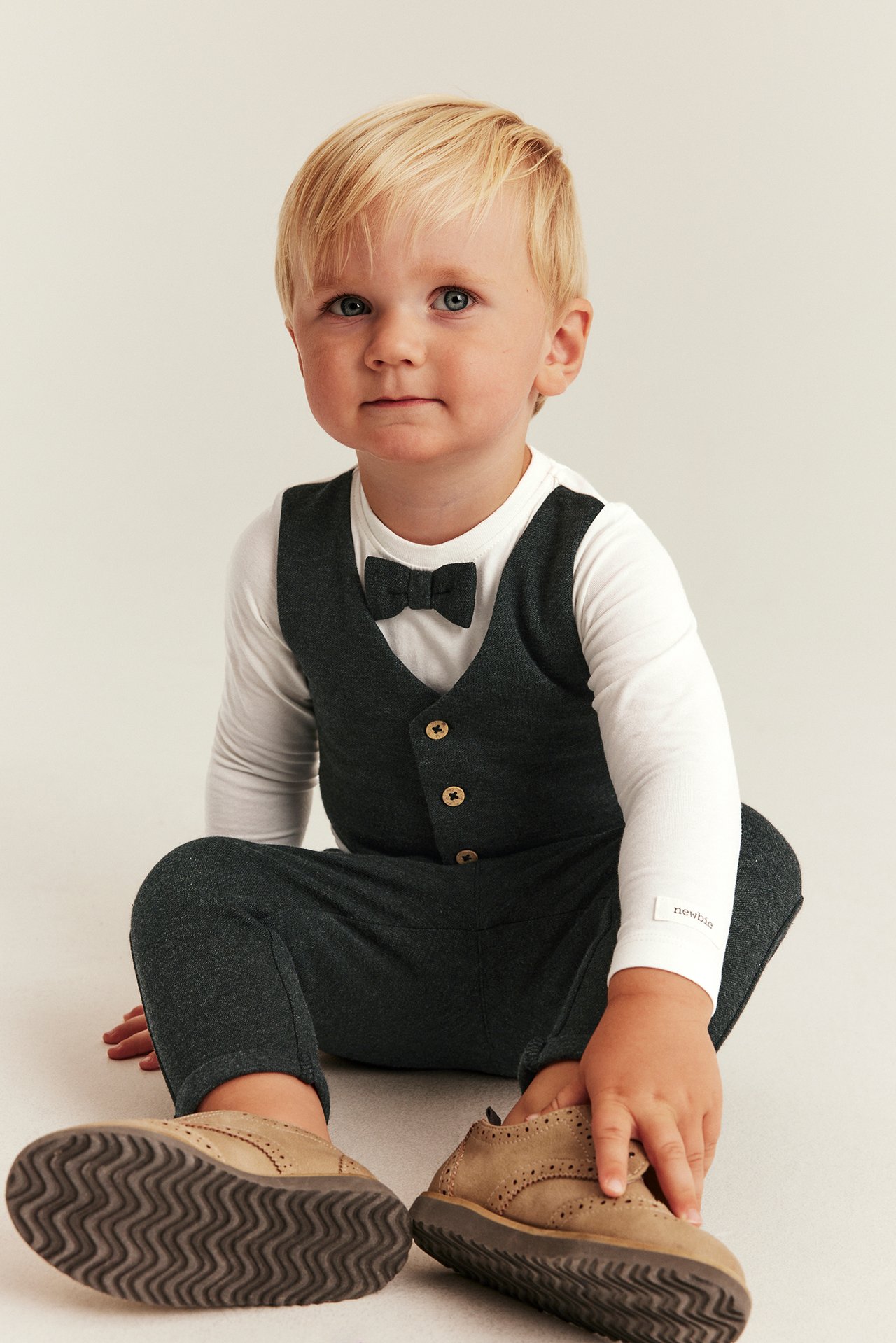 Front view: baby in dark gray long-sleeve romper with mock waistcoat, bow tie, and tan shoes.