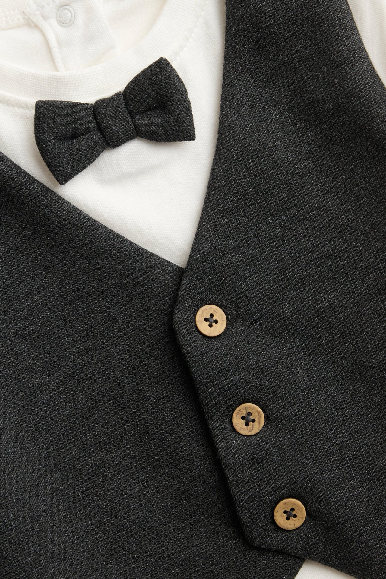 Nahaufnahme, Vorderansicht: Baby-Body in Offwhite mit angenähter dunkelgrauer Weste, Fliege und Holzknöpfen.