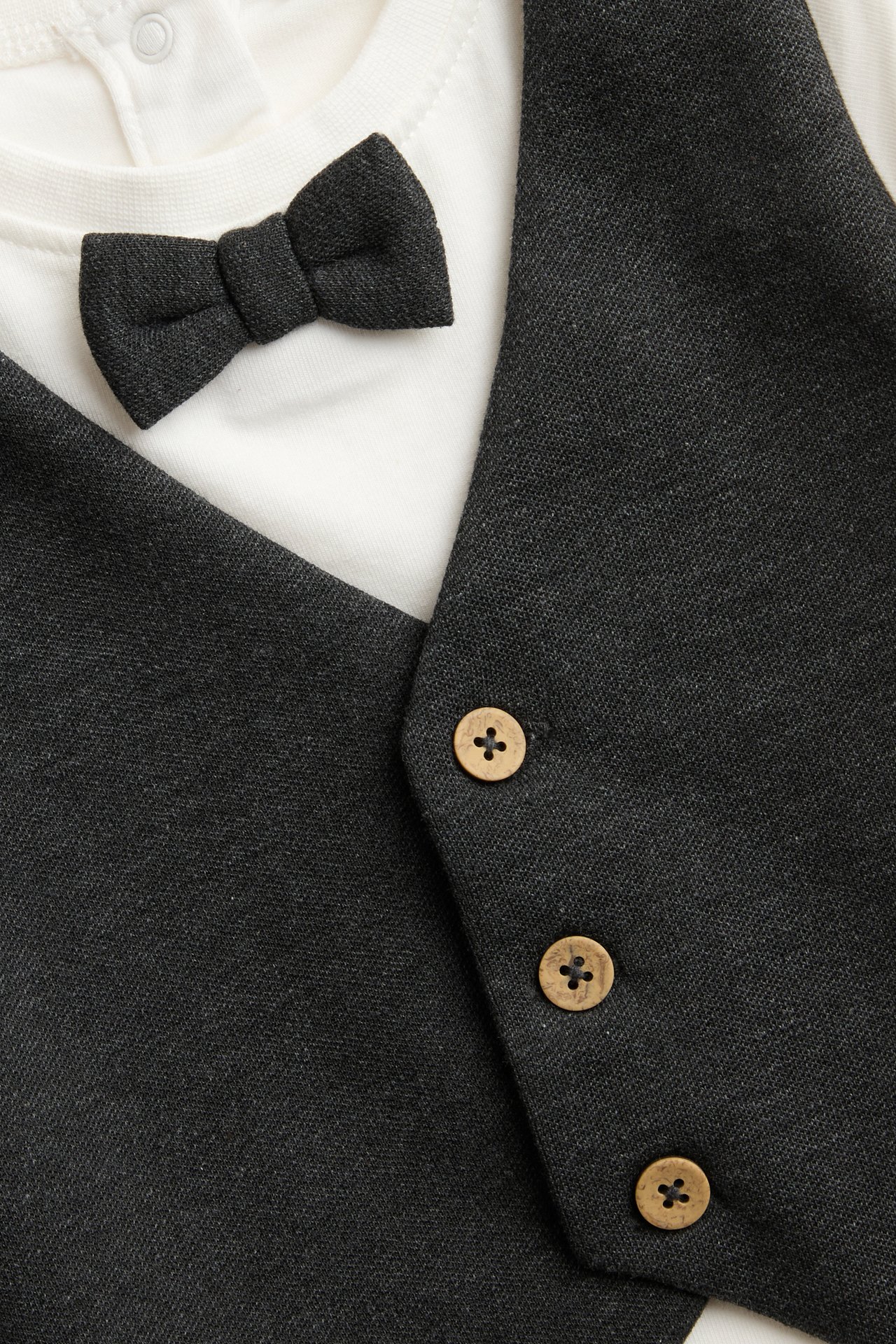 Close-up of a dark gray baby vest with wooden buttons over a white long-sleeve top and bow tie.