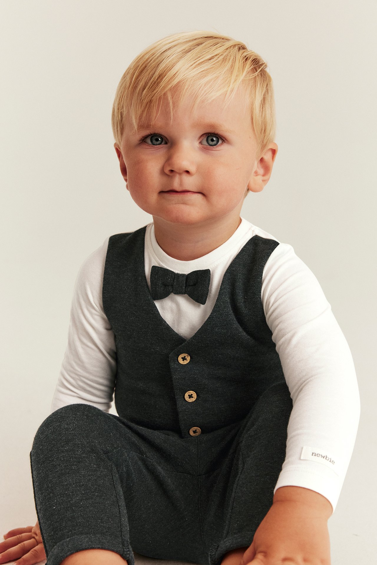 Front view of a baby in a dark gray waistcoat, pants, white long-sleeve top, and dark gray bow tie.