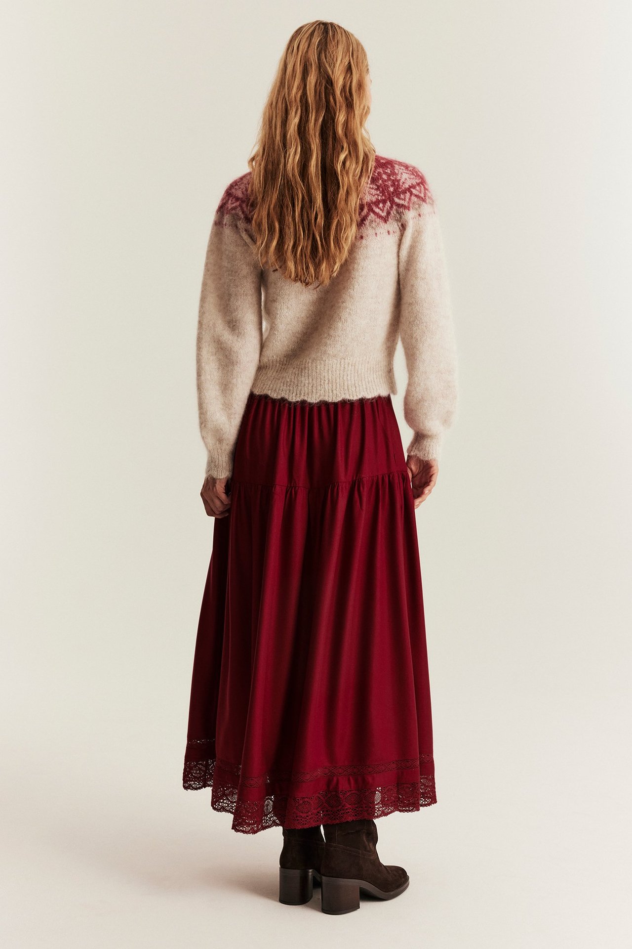 Back view of a woman wearing a long red viscose skirt with lace hem, a patterned sweater, and brown boots.