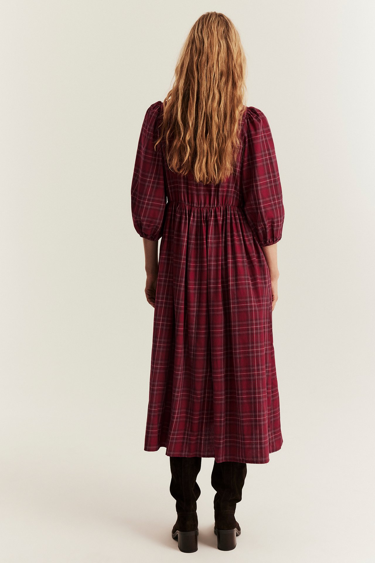 Rückenansicht einer Frau in rotem, kariertem Baumwollkleid mit Puffärmeln und schwarzen Stiefeln.