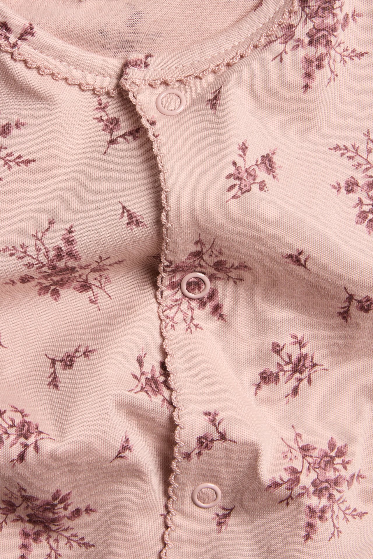 Close-up front view of a pink cotton baby pajama with tone-on-tone floral pattern, snap buttons, and picot trim.