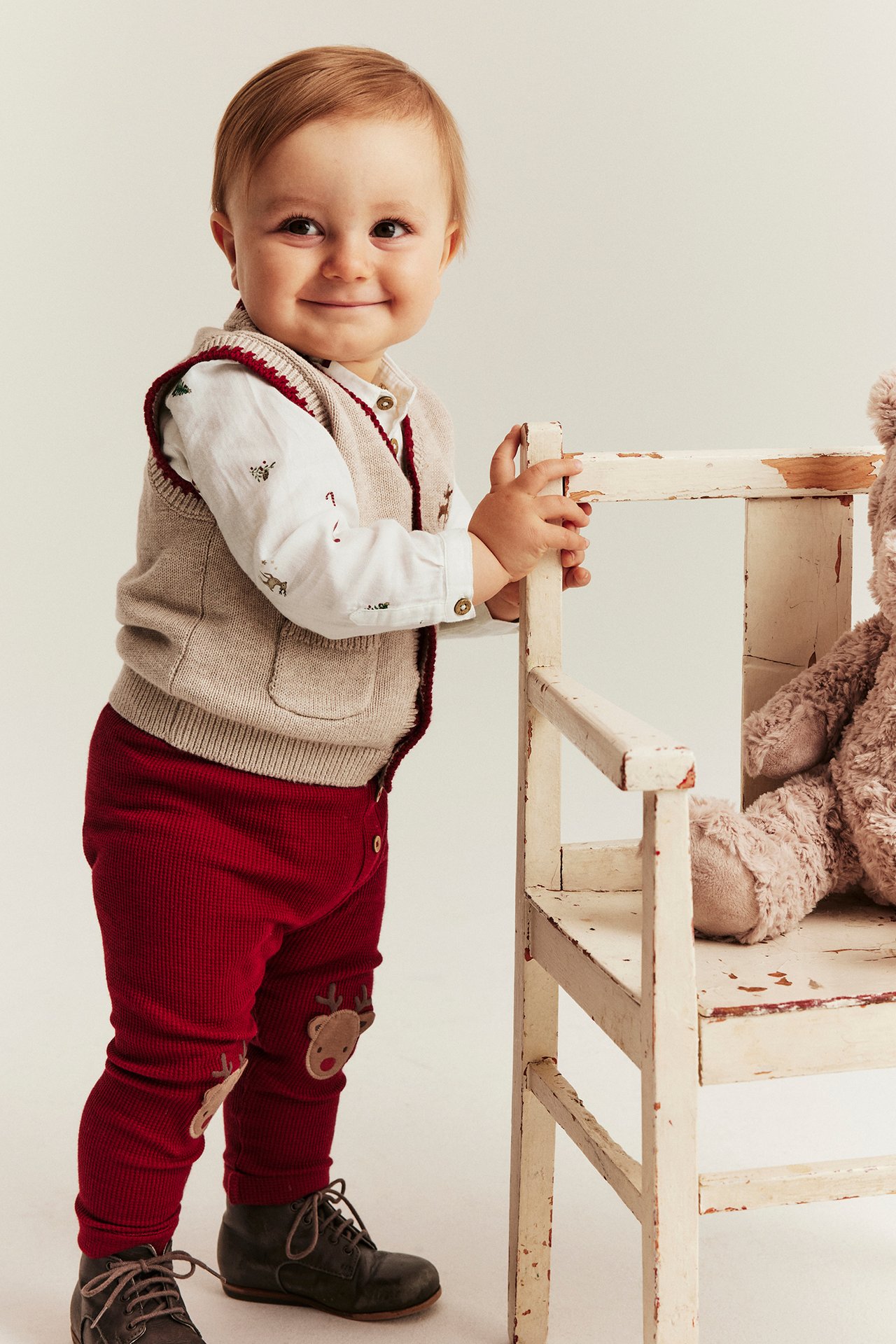 Baby holding chair, wearing off-white animal print bodysuit, beige vest, red reindeer leggings, dark boots.