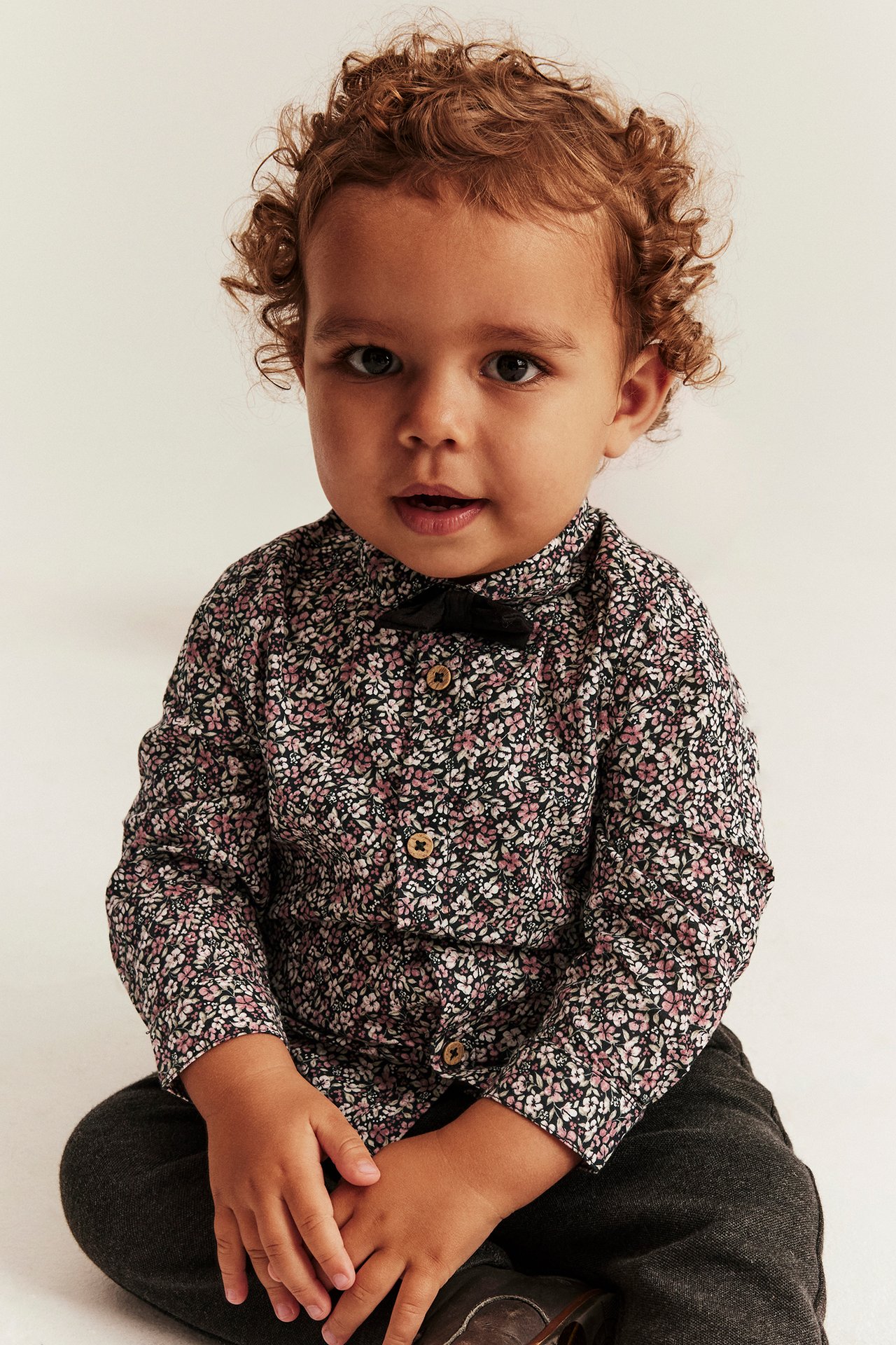 Front view of a baby wearing a black floral shirt with a black bow tie and dark grey trousers.