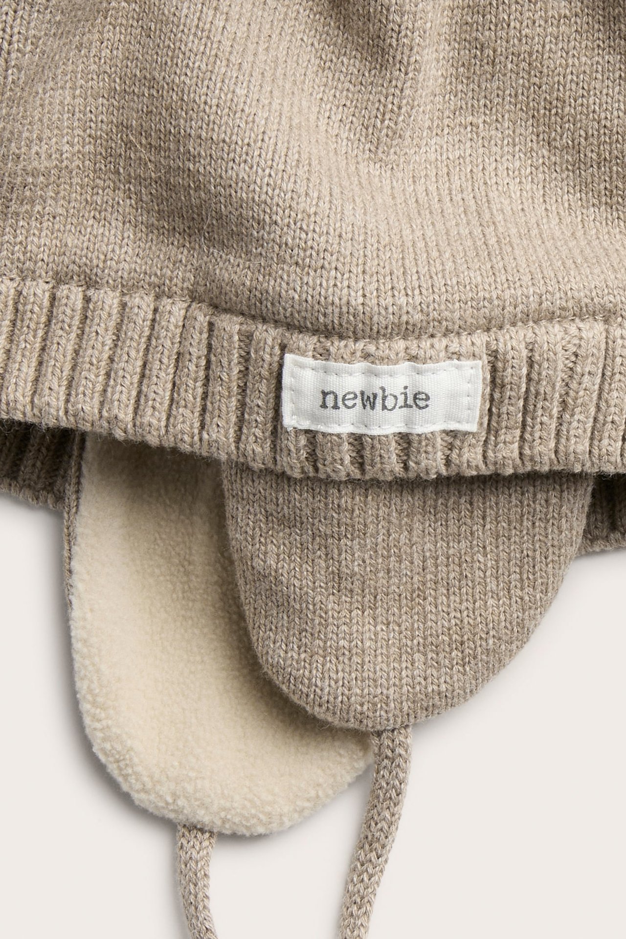 Nahaufnahme einer hellbraunen gestrickten Babymütze mit Ohrenklappen, Bindebändern und cremefarbenem Fleecefutter.
