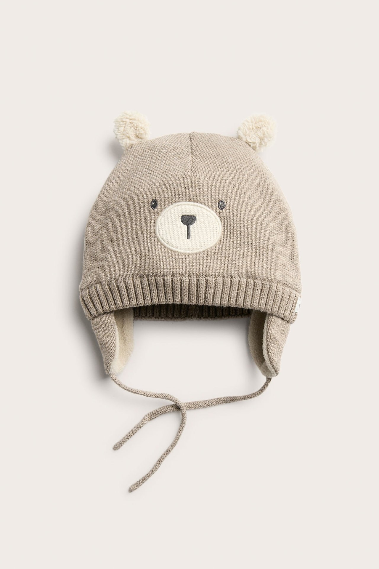 Vorderansicht einer Baby-Mütze in Beige-Melange mit Bärengesicht, Bommelohren, Ohrenklappen und Bindebändern.