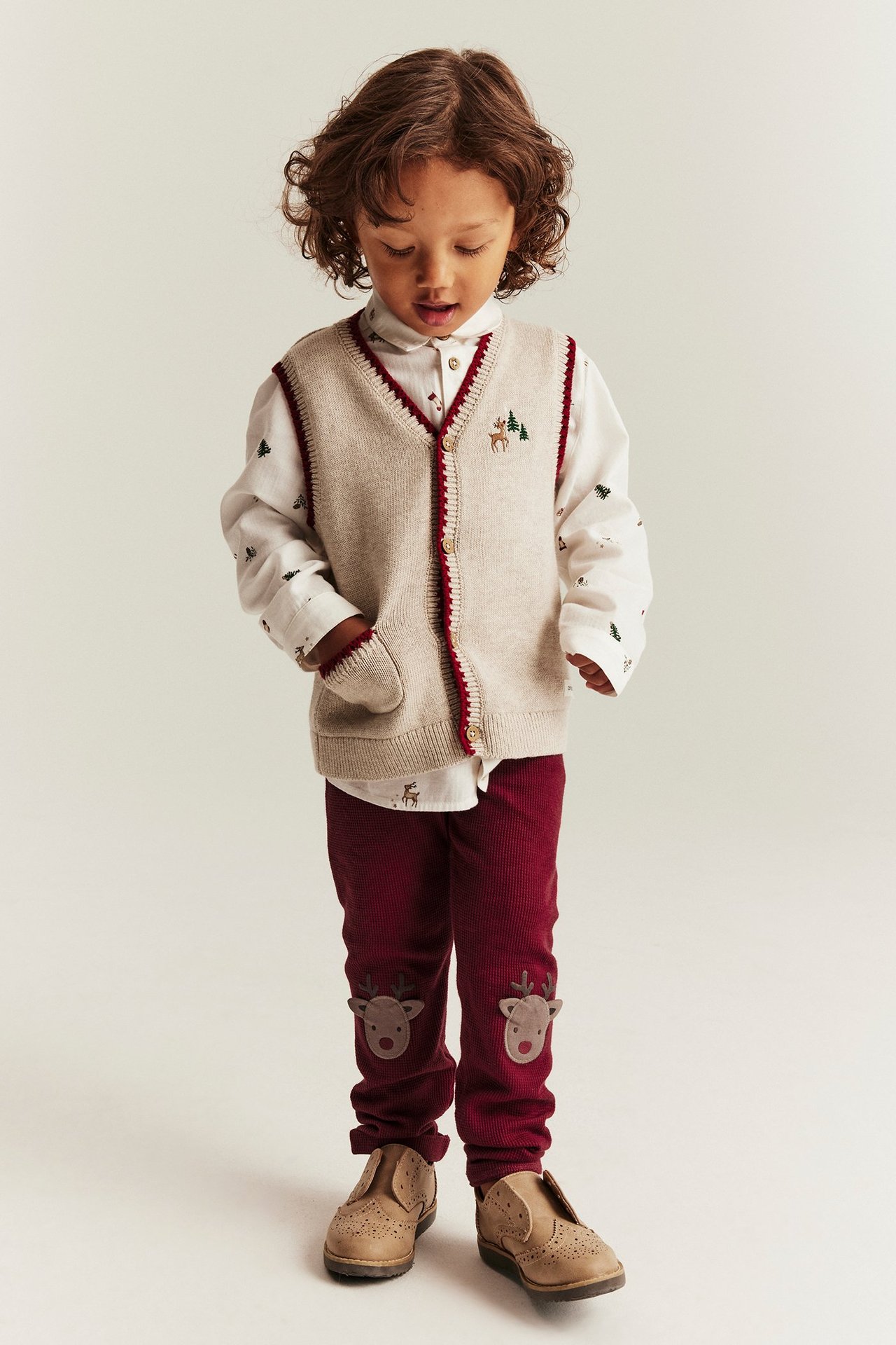 Kid in red reindeer leggings, beige vest with buttons, white shirt, beige shoes.