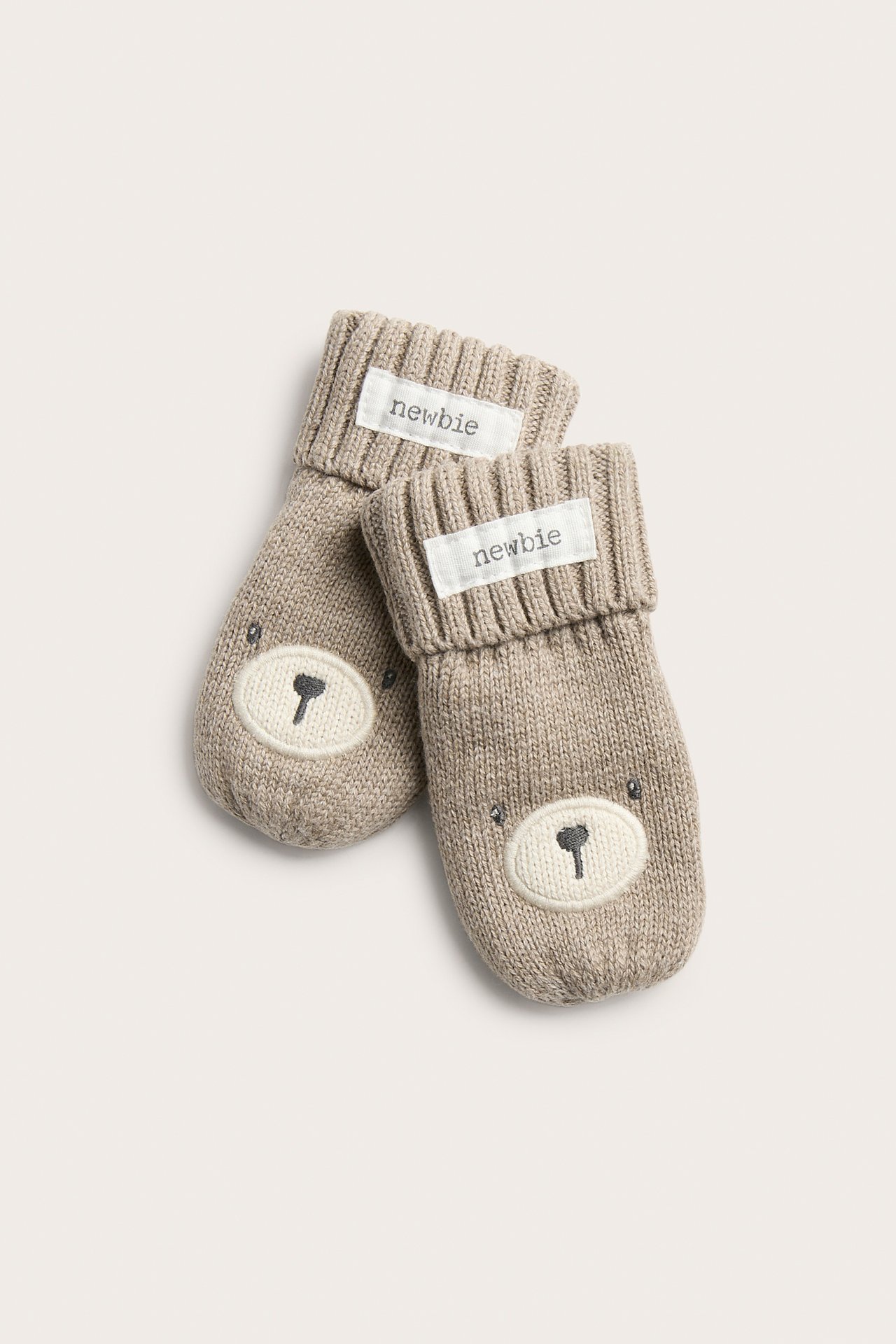 Von oben, zwei hellbraun melierte gestrickte Baby-Fäustlinge mit Bärengesichtern und gerippten Bündchen.