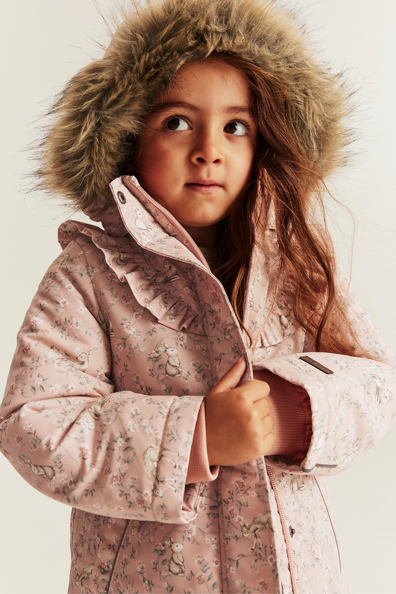 Vorderansicht: Kind in rosa wattierter Blumenjacke mit Hasenprint, Fellkapuze und Rüschenleiste.