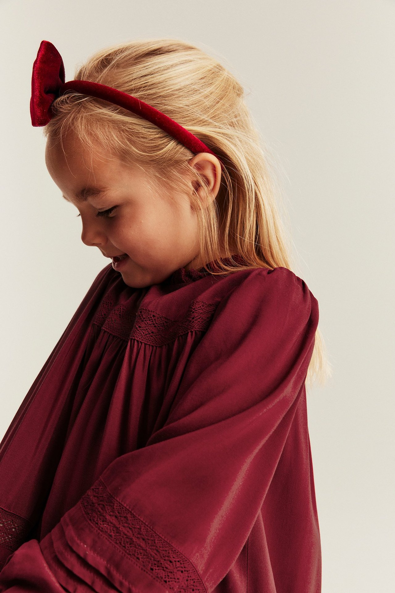 Side view of a kid wearing a red viscose dress with lace details and a red velvet bow headband.