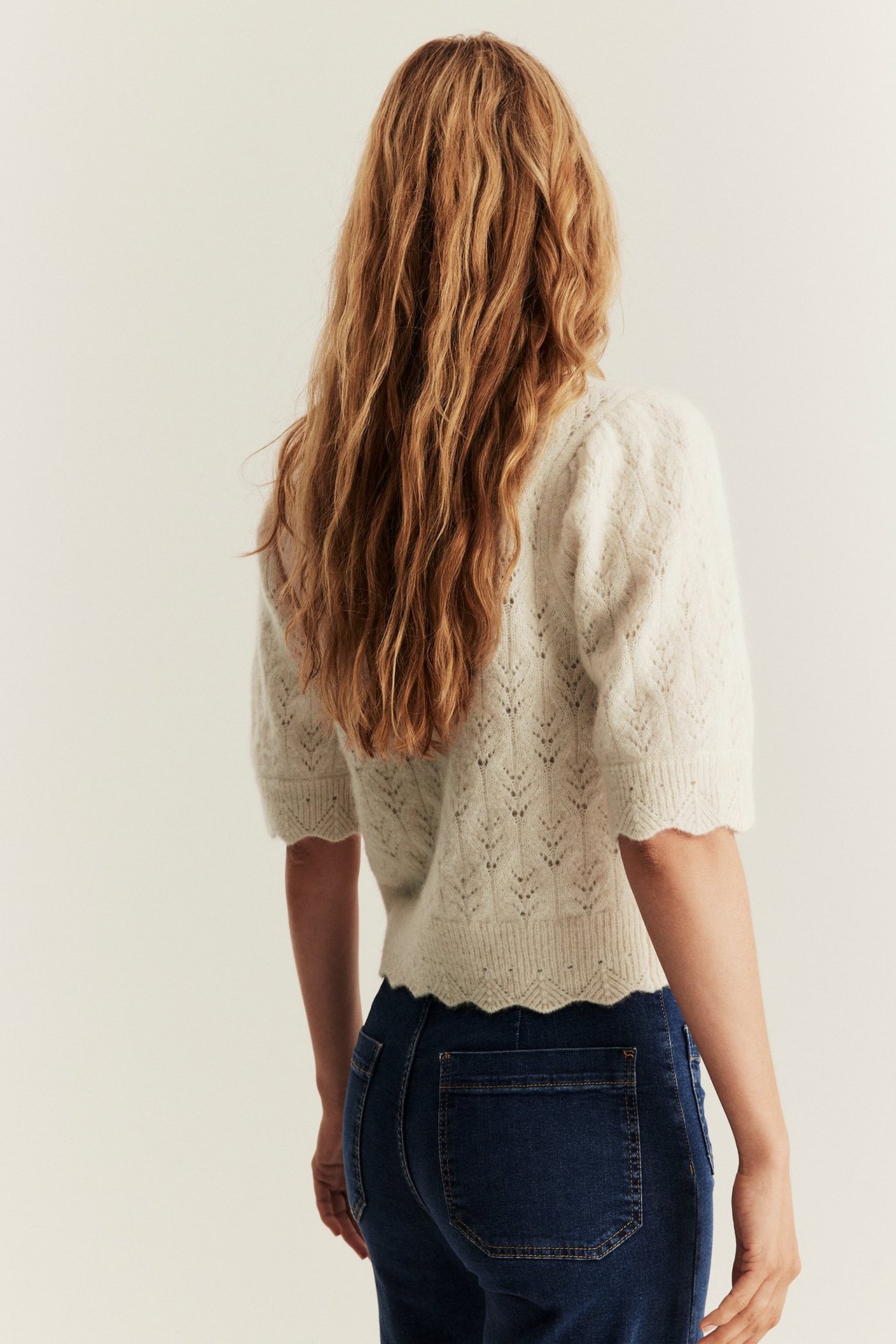 Back view of a woman wearing a light beige patterned knit cardigan with short puff sleeves and dark blue jeans.