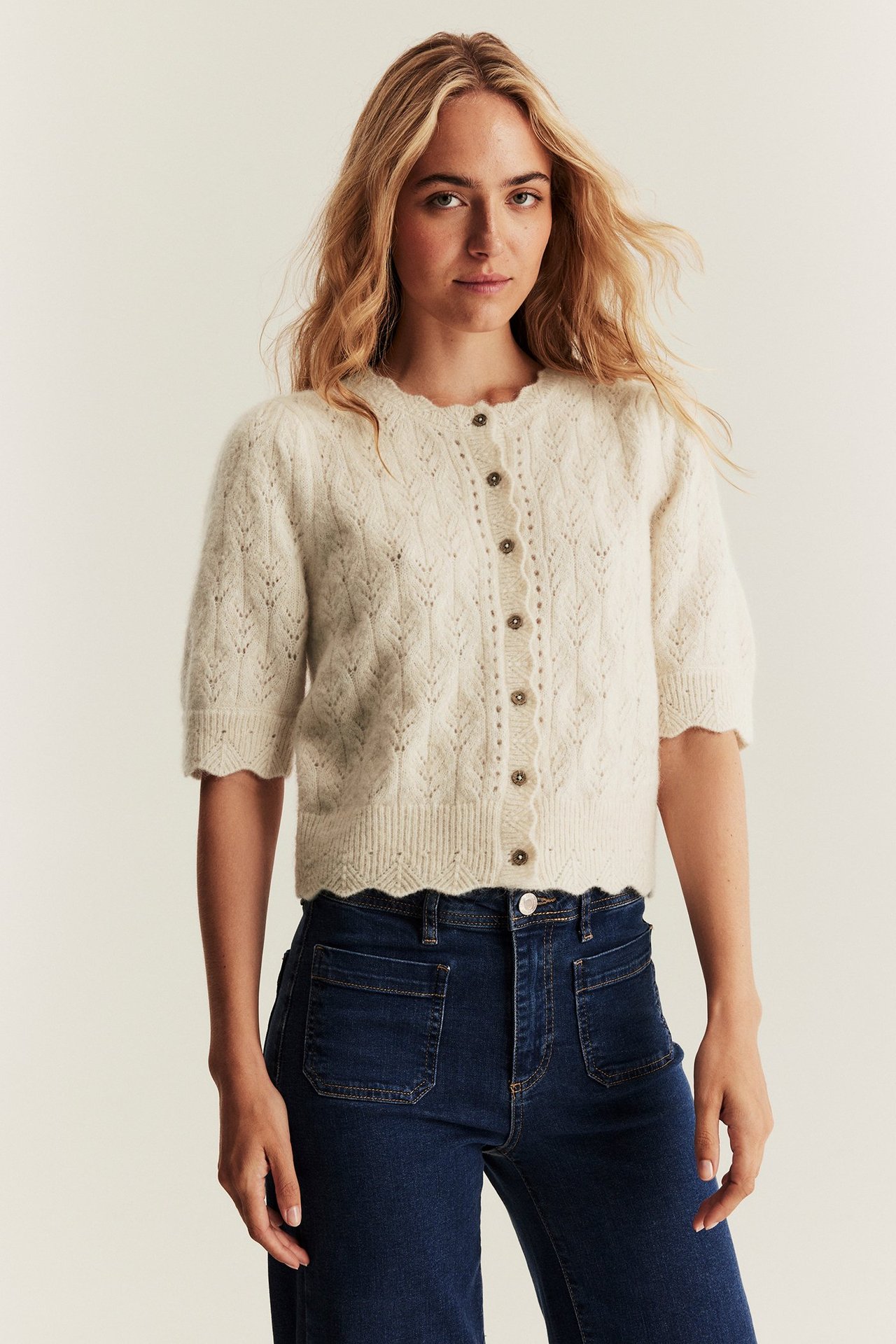 Front view of a woman wearing a light beige patterned knit wool blend cardigan with short puff sleeves and dark blue jeans.