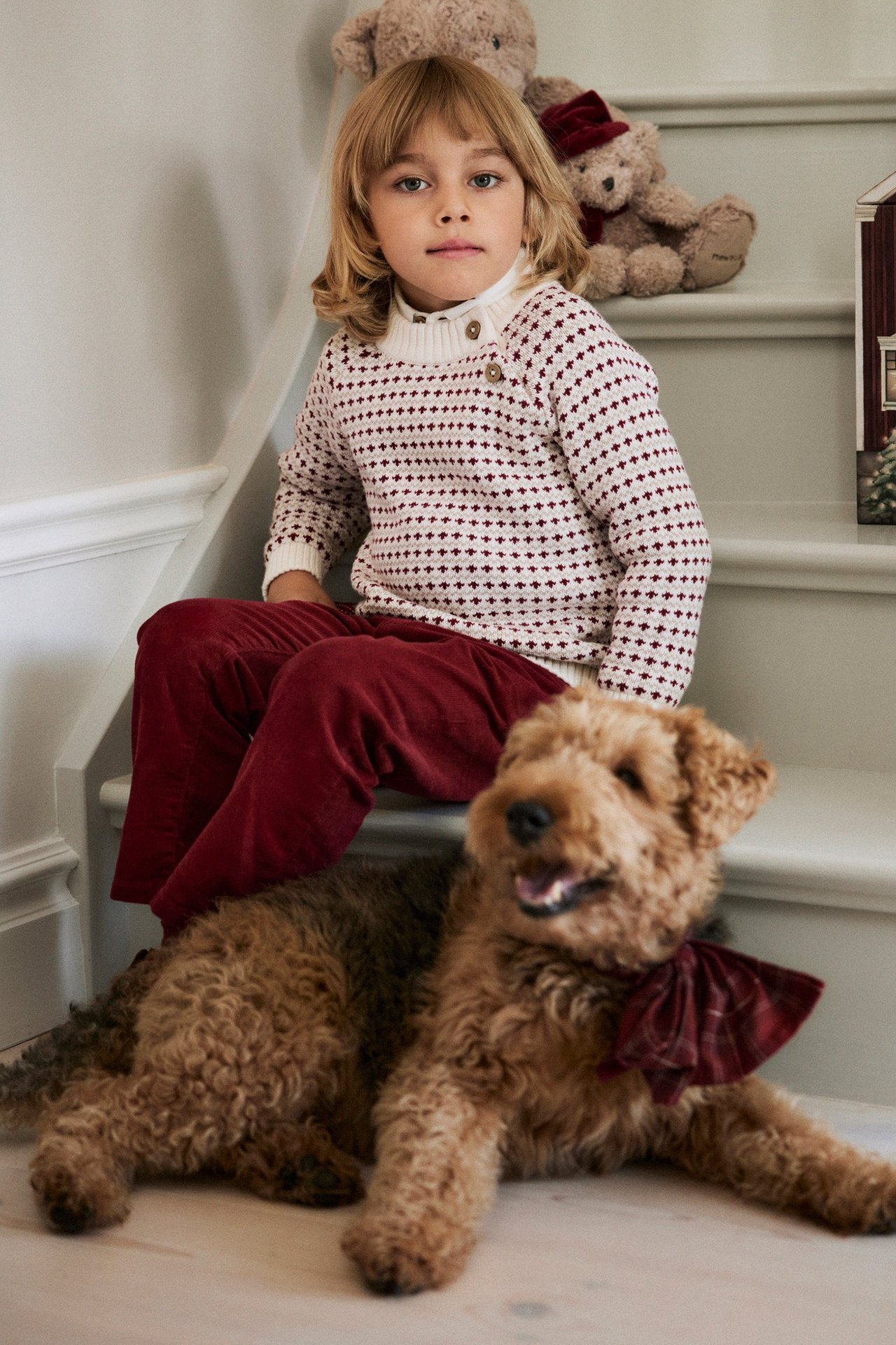 Vorderansicht: Kind trägt einen cremeweißen Jacquard-Strickpullover mit Knöpfen und dunkelrote Hosen.