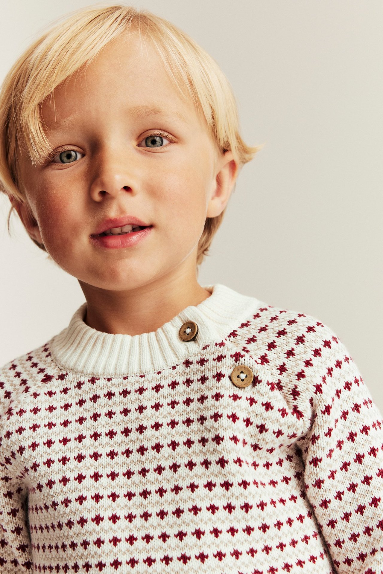Girl in offwhite knitted sweater with red pattern, buttons at shoulder, front view.