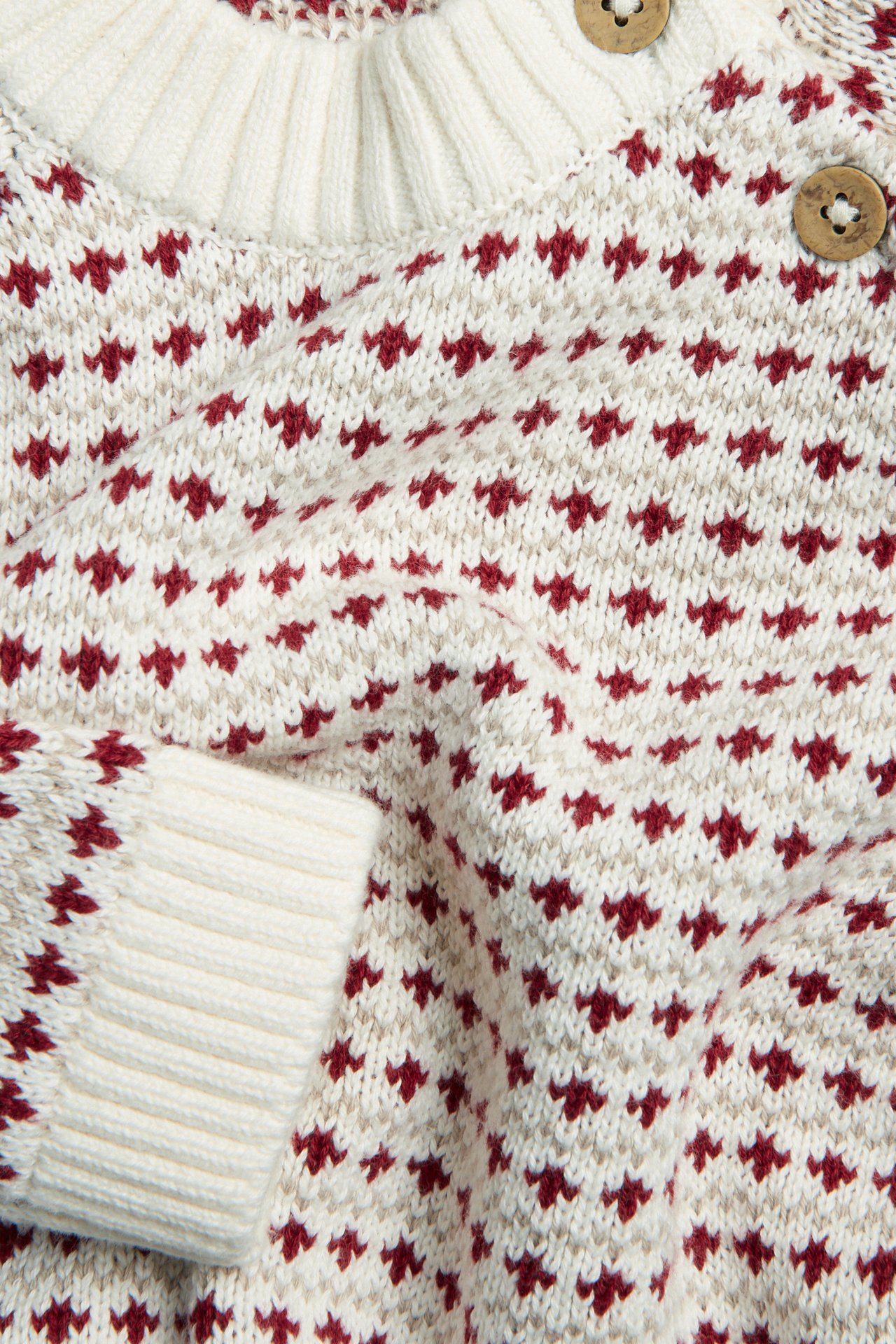 Close-up of an off-white cotton knitted sweater with a red and beige jacquard pattern, ribbed cuffs, and wooden buttons.