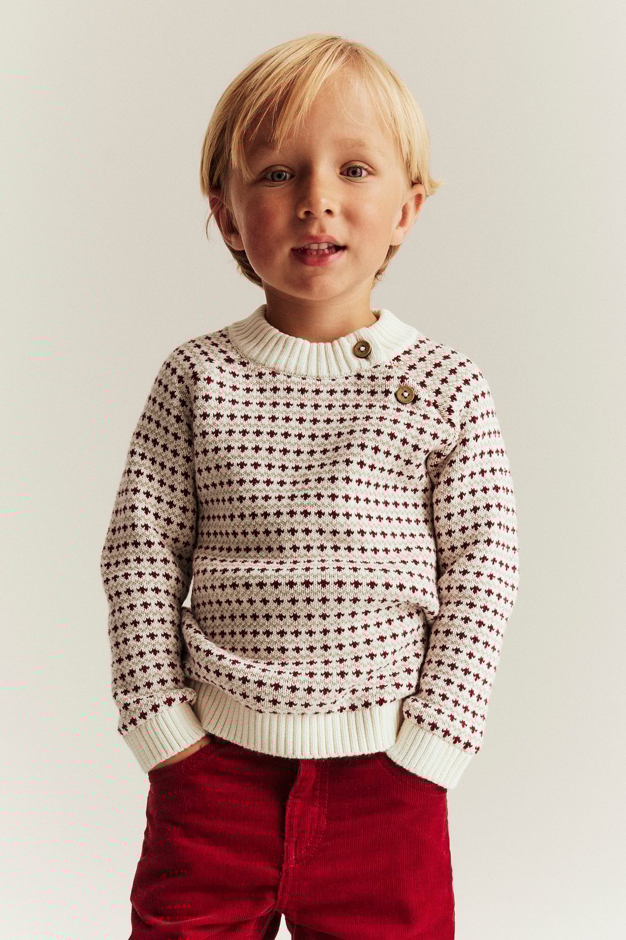 Junge in offwhite Strickpullover mit rotem Jacquardmuster, Knöpfe, rote Hose, Frontansicht.
