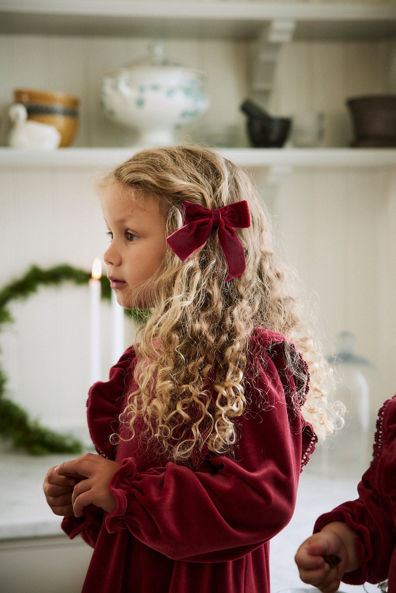 Side view of a kid with a red velvet bow in her hair, wearing a red velvet dress.