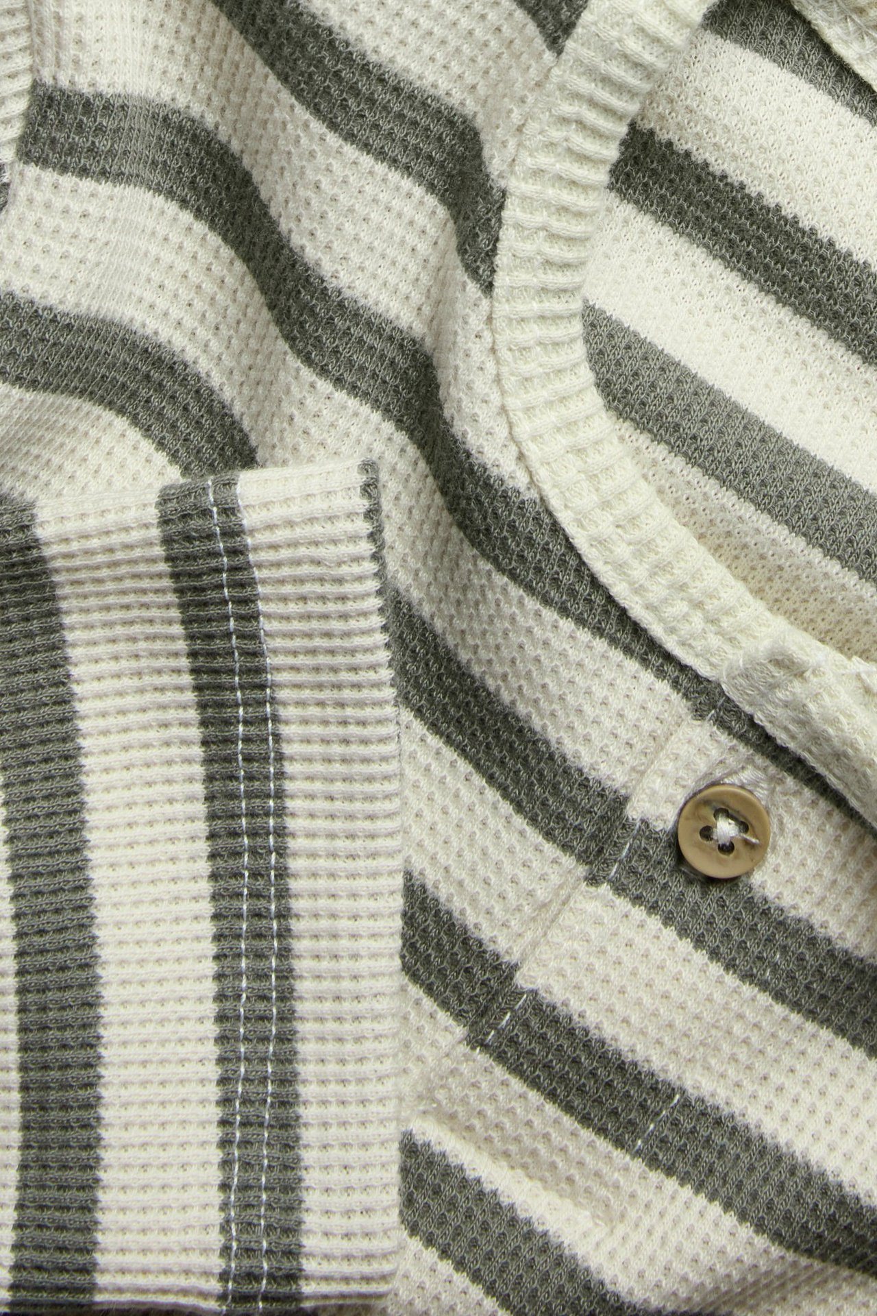 Nahaufnahme: Gestreiftes Waffelstrick-Langarmshirt mit cremeweißen, dunkelgrünen Streifen und einem braunen Knopf.